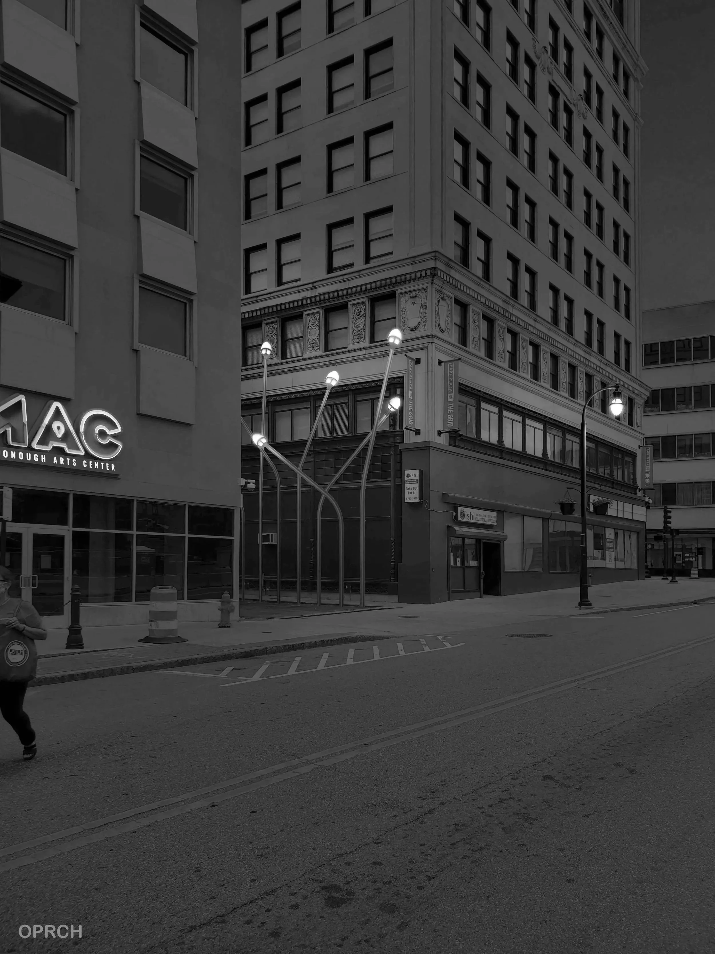 Black and white photo of storefronts and buildings on a city street, with street lamps and a person walking on the sidewalk.