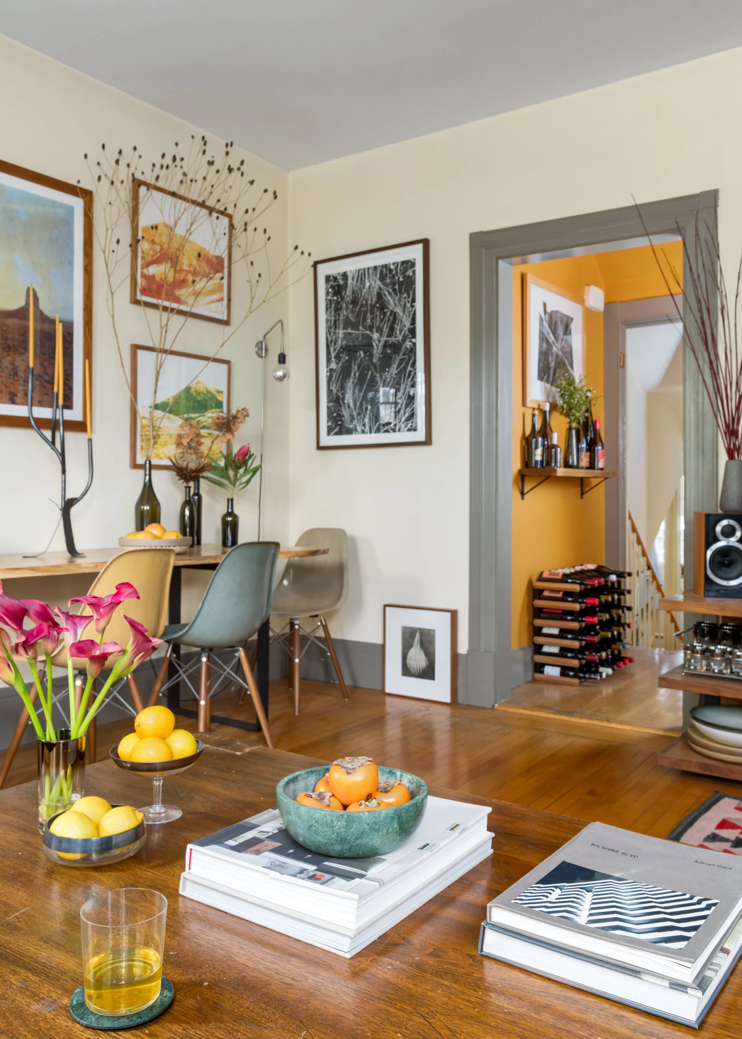 A dining room with a wooden table set with magazines, glass of lemon water, vases with flowers and lemons, and framed artwork on the walls. There's a doorway leading to another room with a wine rack and more artwork.