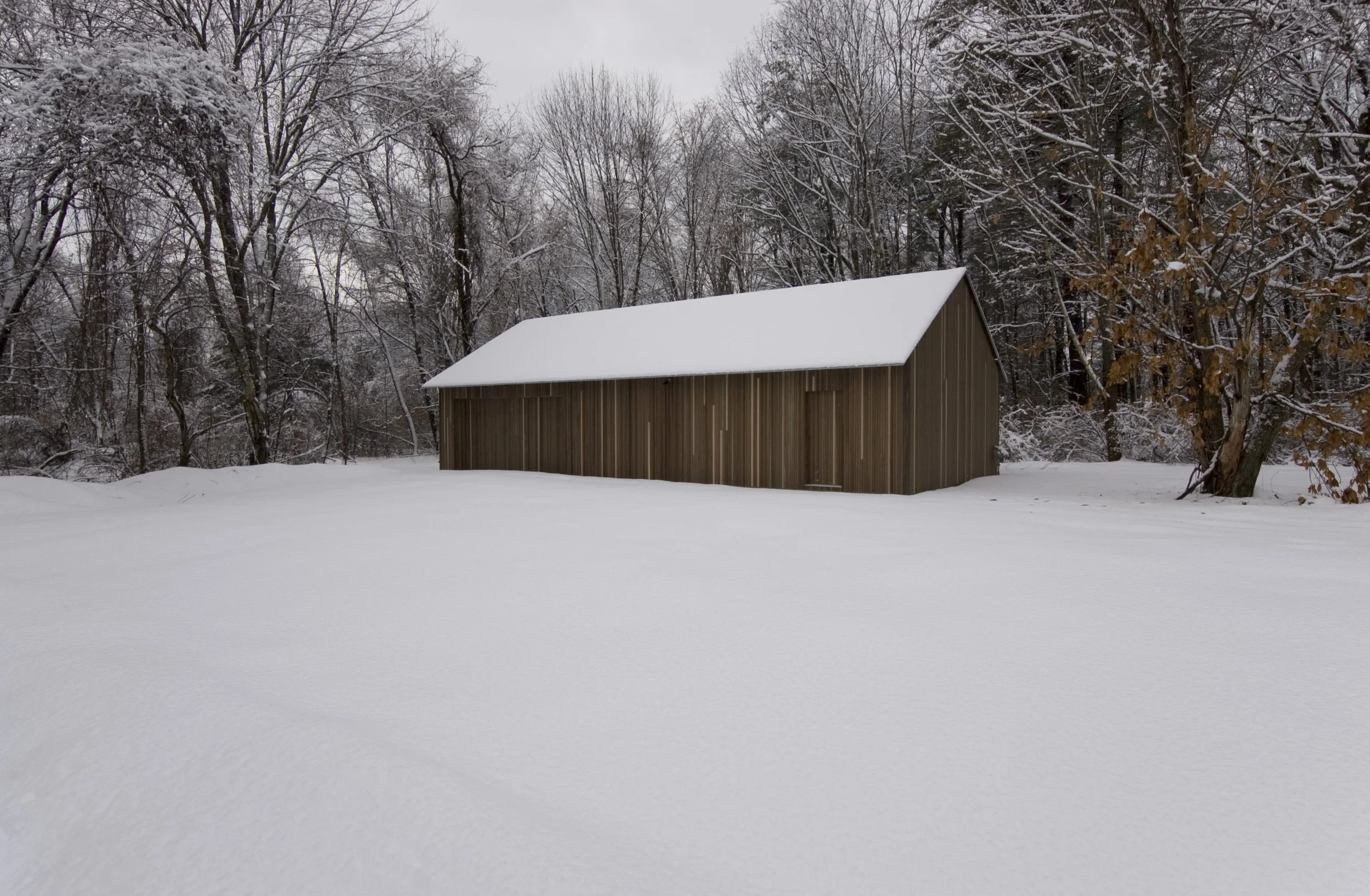 A wooden barn with a snow-covered roof in a snowy landscape surrounded by trees with snow on their branches.
