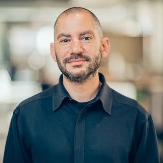 Portrait of a man with a beard and short hair, wearing a dark button-up shirt, standing indoors.