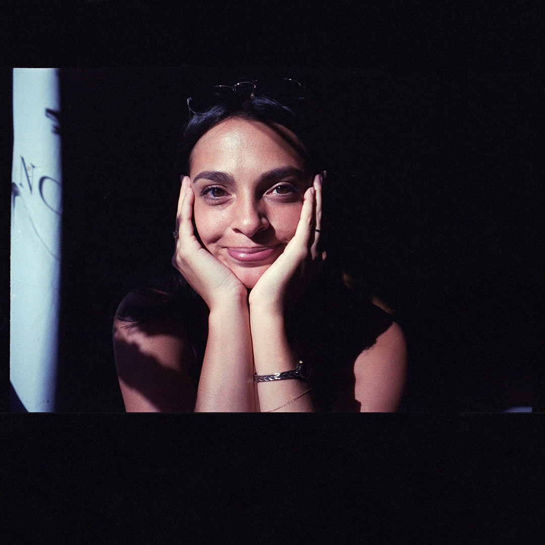 A young woman with dark hair and a gentle smile, resting her face in her hands, with light shining on her face.