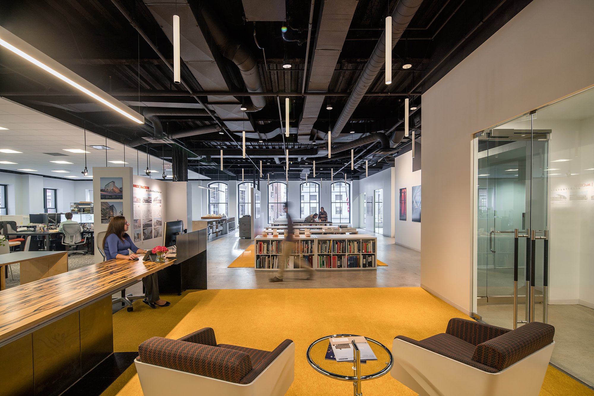 Interior of an architecture office with white walls, large windows, and black ceiling with exposed pipes and hanging lights. A woman is sitting at a wooden reception desk working on a computer, with office furniture and people in the background.