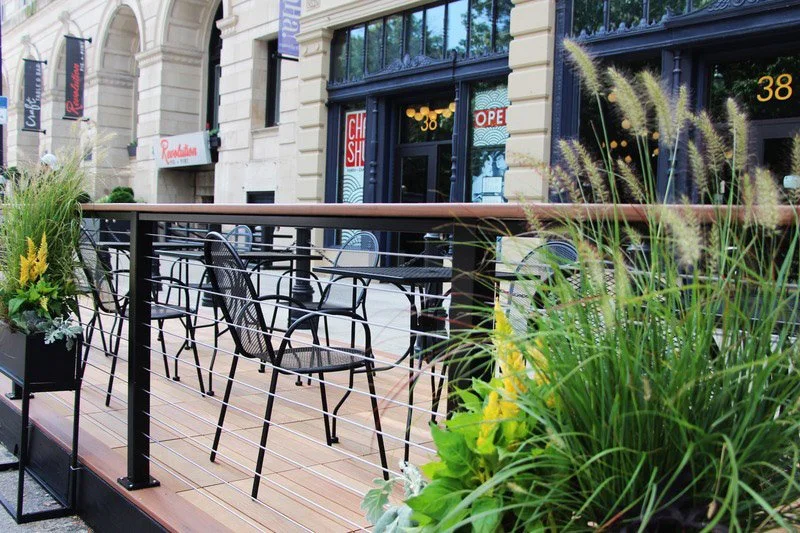 Outdoor patio seating area with black metal chairs and tables, surrounded by planters with tall grasses and green plants, in front of a building in Worcester, MA.