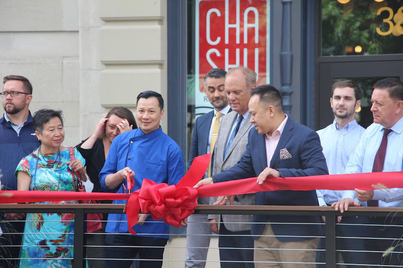 People attending a ribbon-cutting ceremony outside a storefront with a sign that says 'Cha Shu' in red. One man in a dark suit is cutting the ribbon.