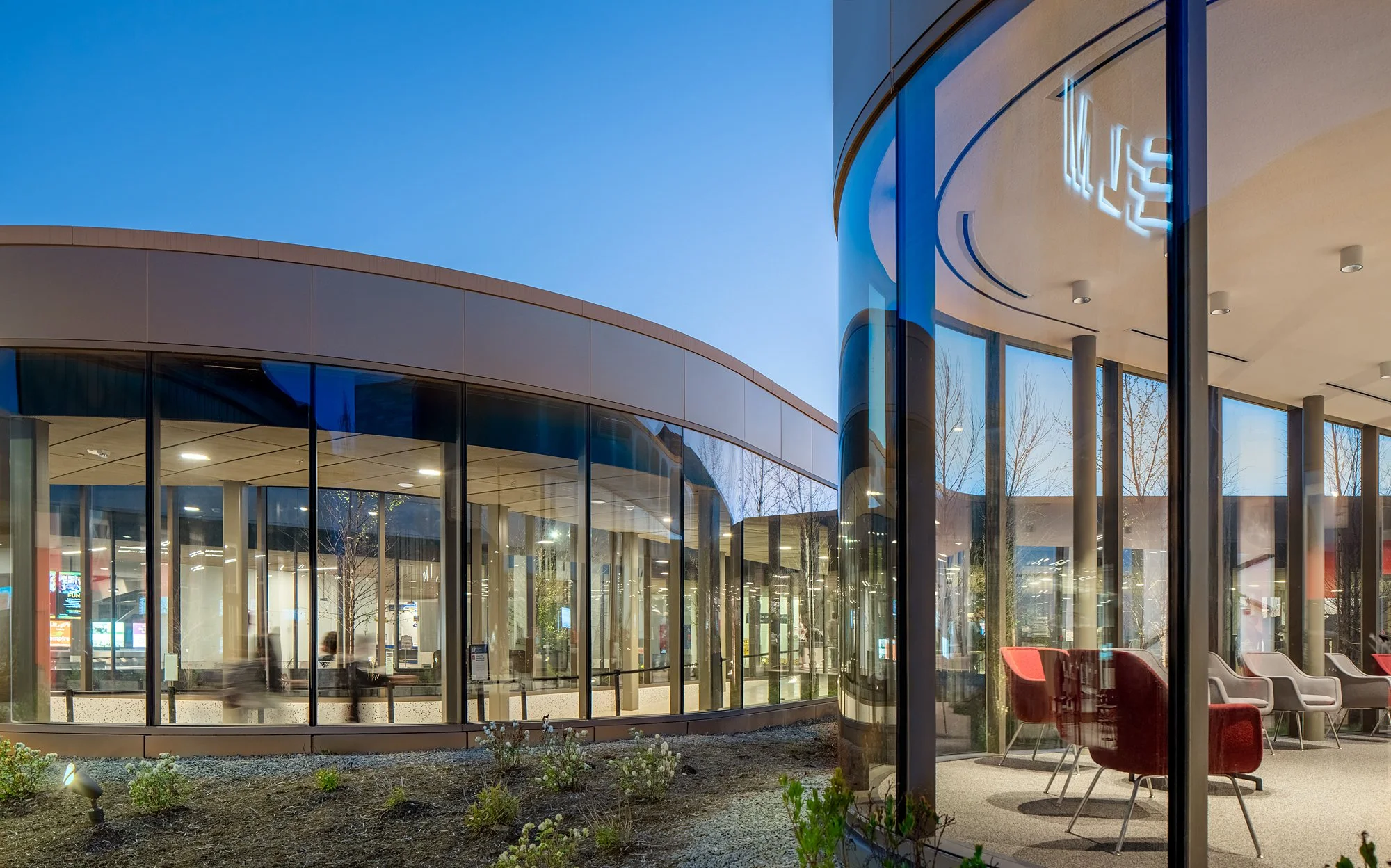 Exterior view of a modern building with curved glass walls and a clear blue sky, showing the interior seating area through the glass.