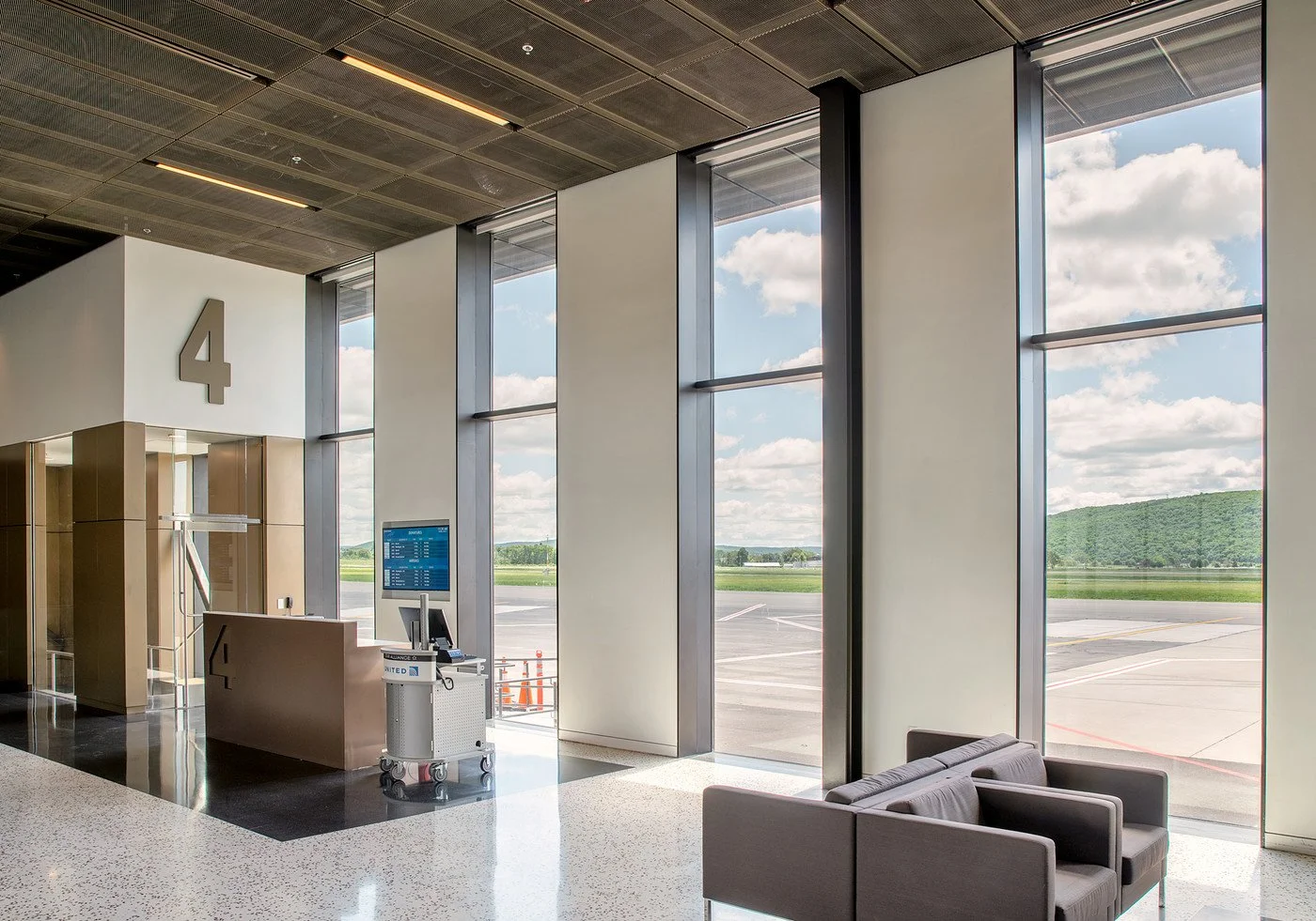 A modern airport terminal with large windows showing the runway and sky outside. There are check-in counters, a seating area with chairs, and a digital flight information display.