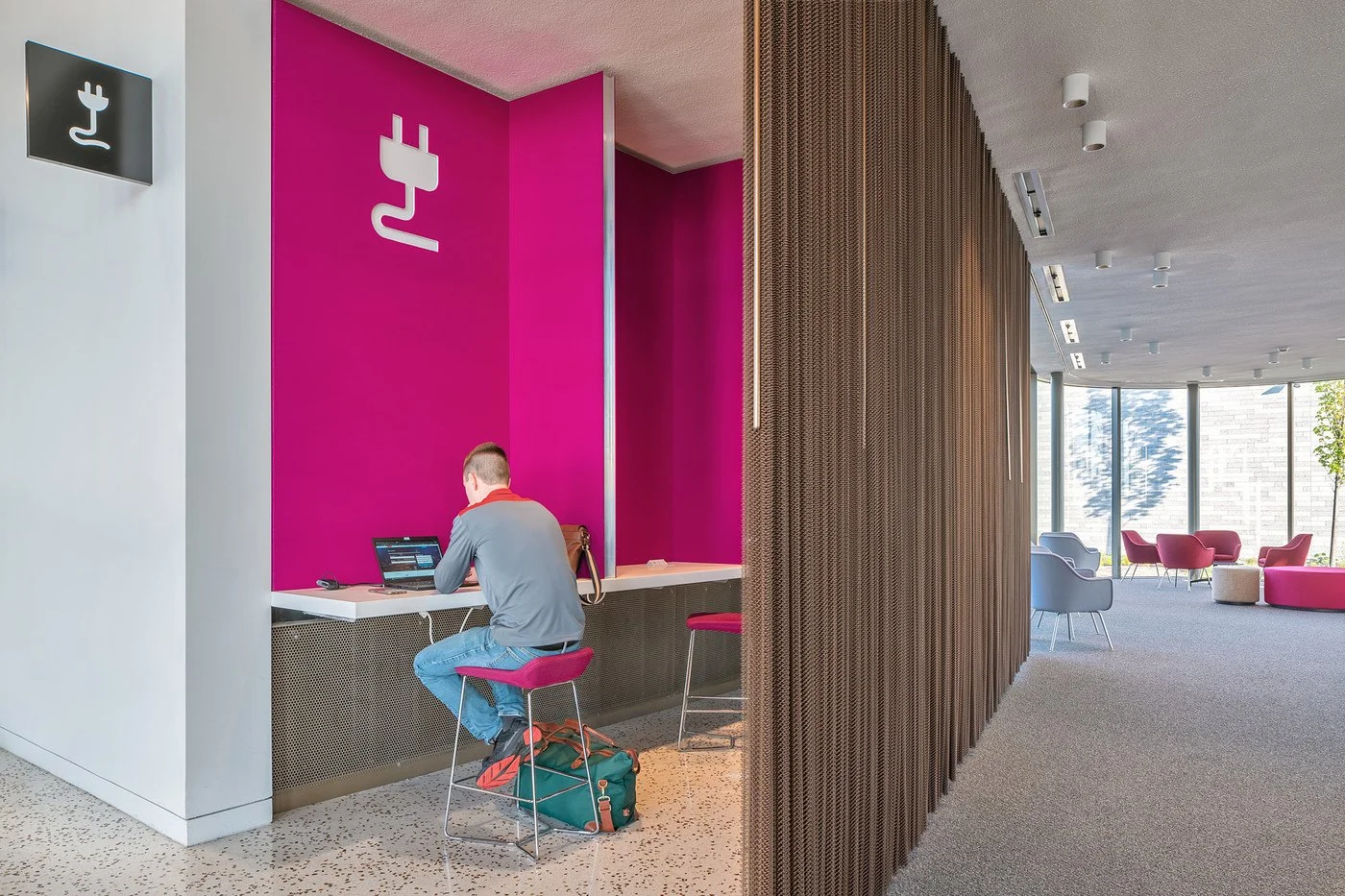 A man sitting at a counter using a laptop in a modern, brightly colored lobby or lounge area with pink and gray furniture and floor-to-ceiling windows.