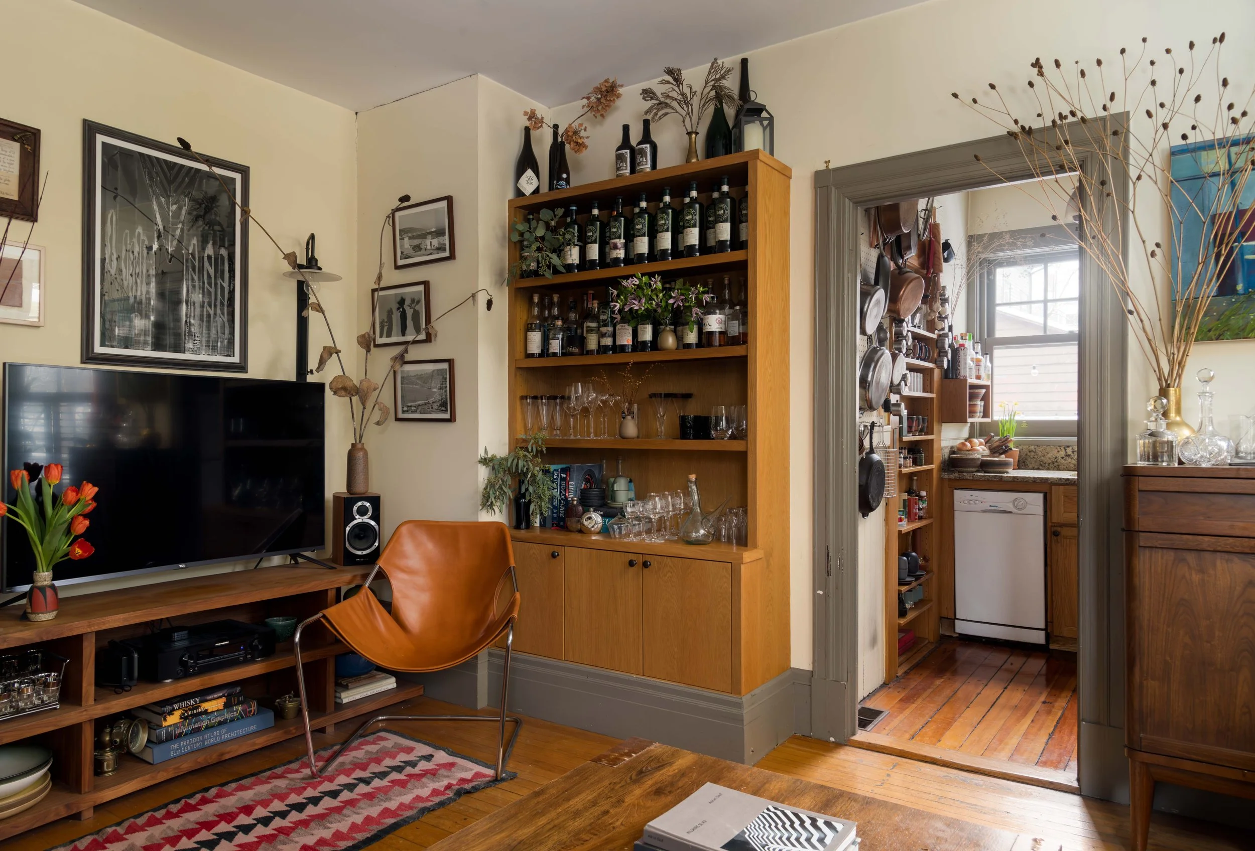 Living room with wall-mounted TV, framed art, decorative plants, and wooden shelves holding wine bottles, glasses, and decorative items, with a doorway leading to a kitchen area.