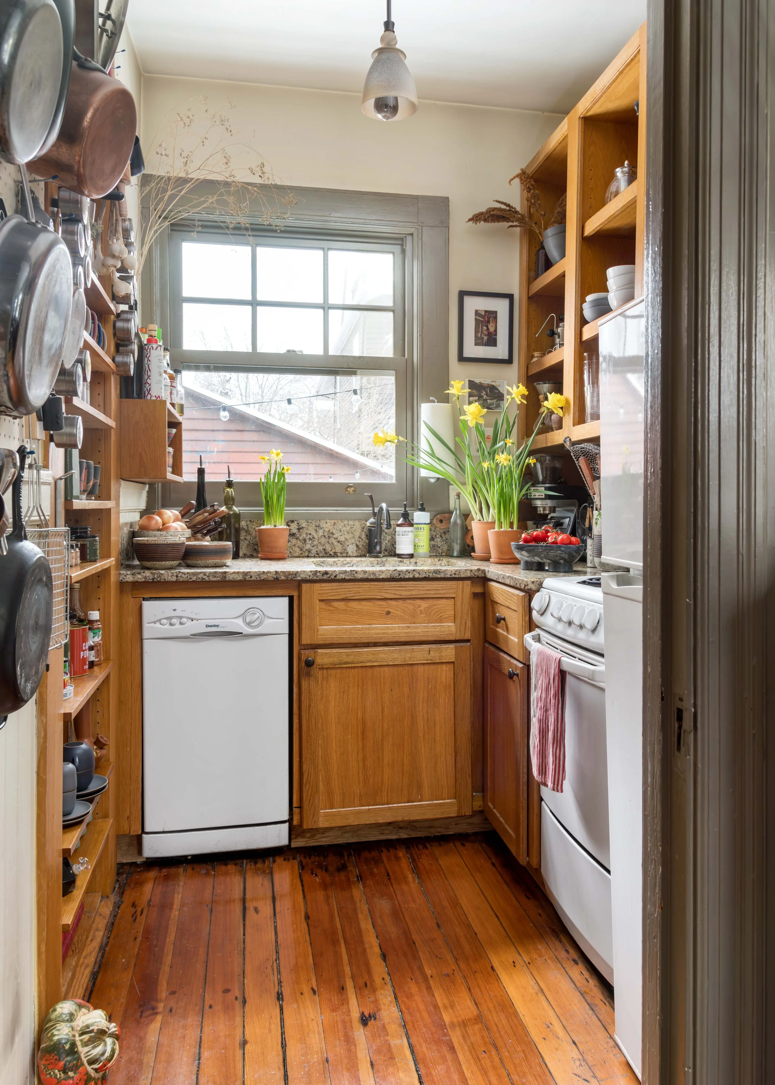 A small kitchen with wooden cabinetry, a white dishwasher, a white stove, and a window above the sink. Potted daffodils and other items are on the counter; hanging pots are on the left wall. The floor is wooden, and light from the window illuminates 