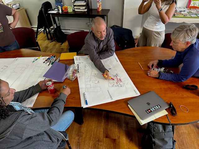 Group of five people gathered around a large wooden table, engaged in a discussion of the project, with plans, markers, and a laptop on the table.