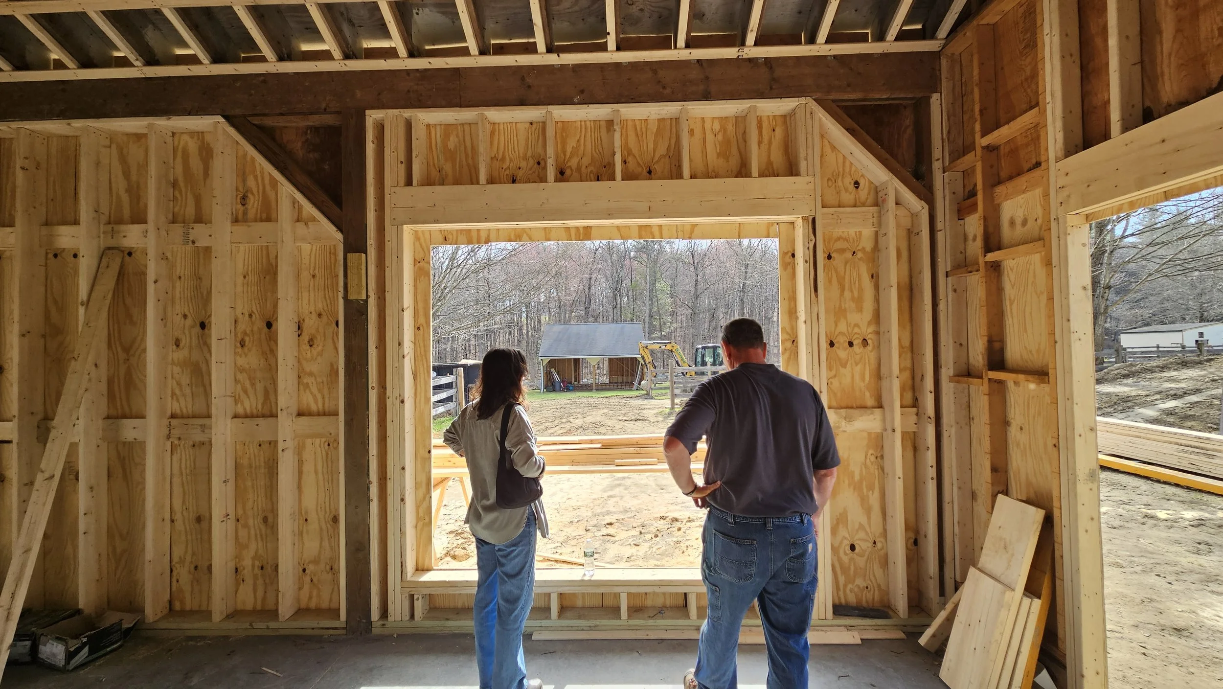 Two people stand inside a wooden frame of a building under construction, looking outside at a yard with trees, a shed, and construction equipment.
