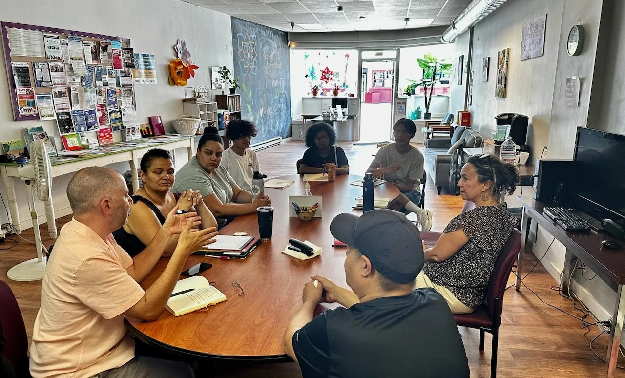A group of eight people sitting around a large oval meeting table in a community room, engaged in a discussion or meeting.