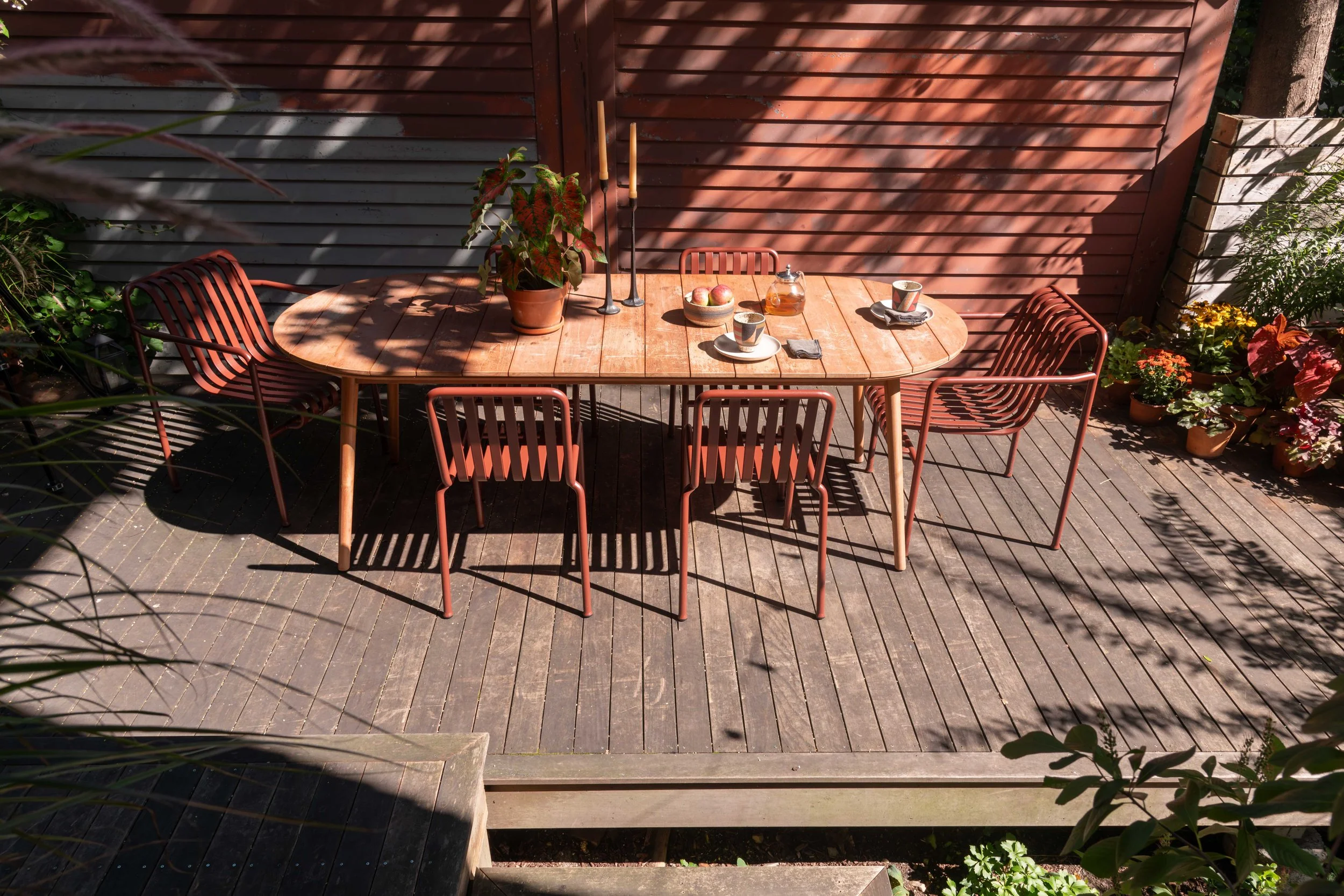 A round wooden outdoor table set on a wooden deck, with six red metal chairs arranged around it. The table has a small bowl of apples, a glass teapot, three teacups, candles, and some napkins. Bright sunlight creates shadows from nearby trees and sla