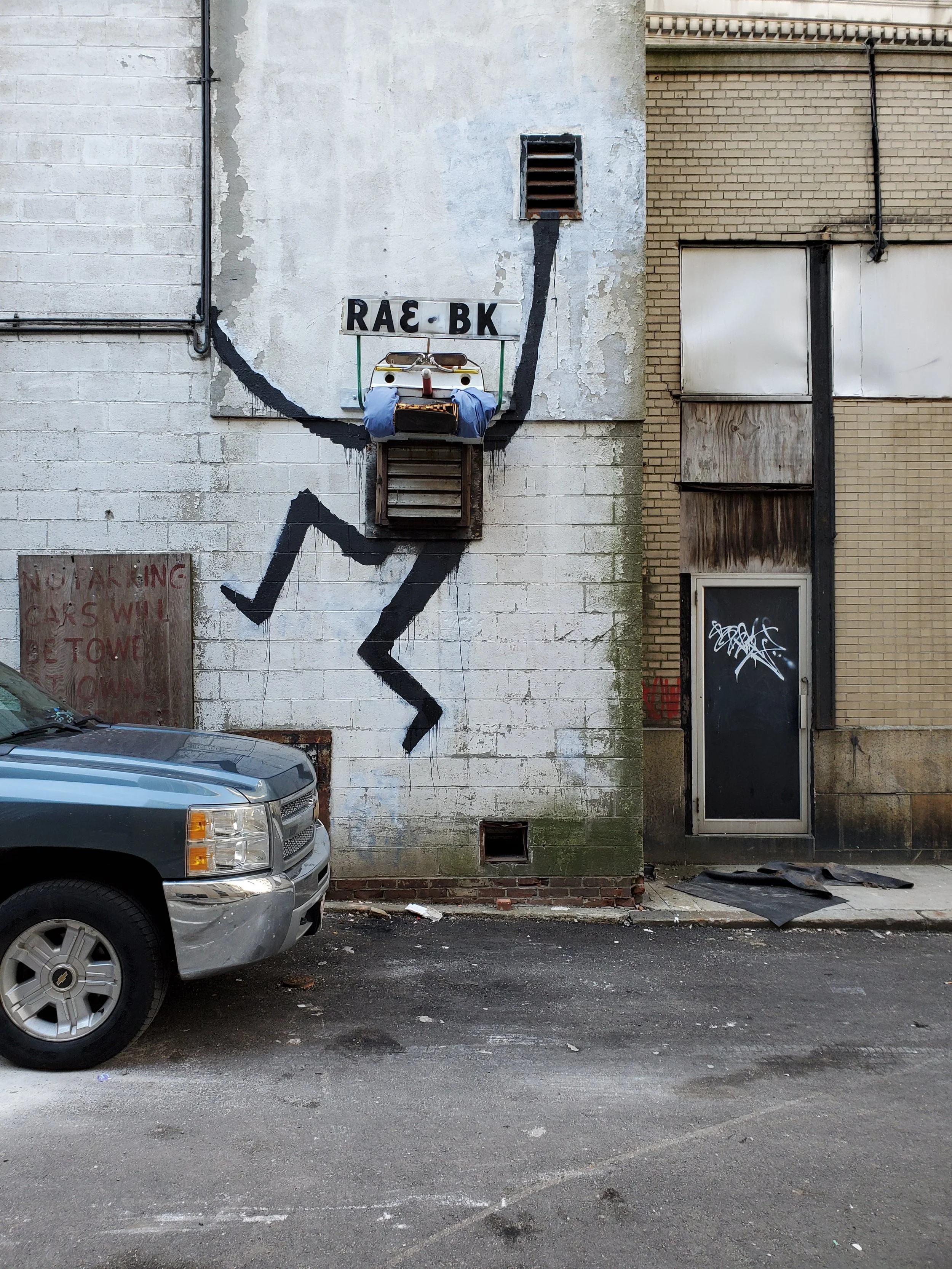 Urban alley wall with graffiti, old vent, and part of a parked silver vehicle in the foreground. The wall has a painted black figure and a corrupted sign with the letters 'RAE·BK'.