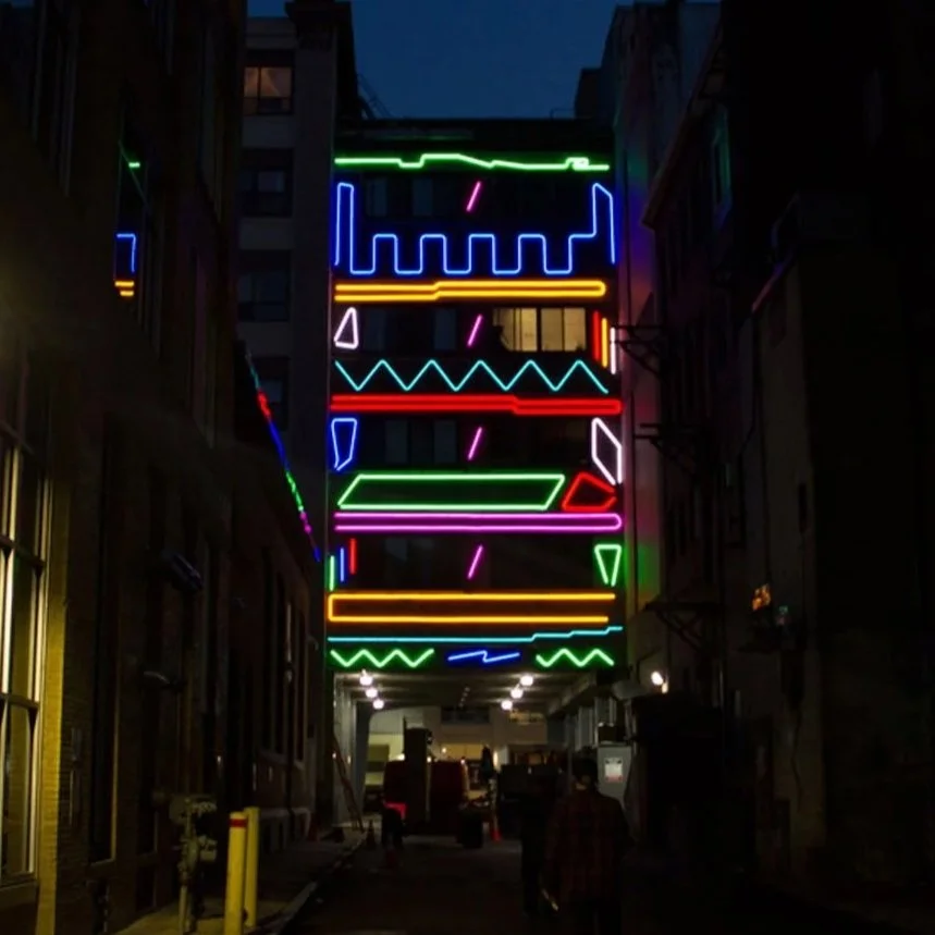Colorful neon geometric shapes and lines hanging on an alleyway at night.
