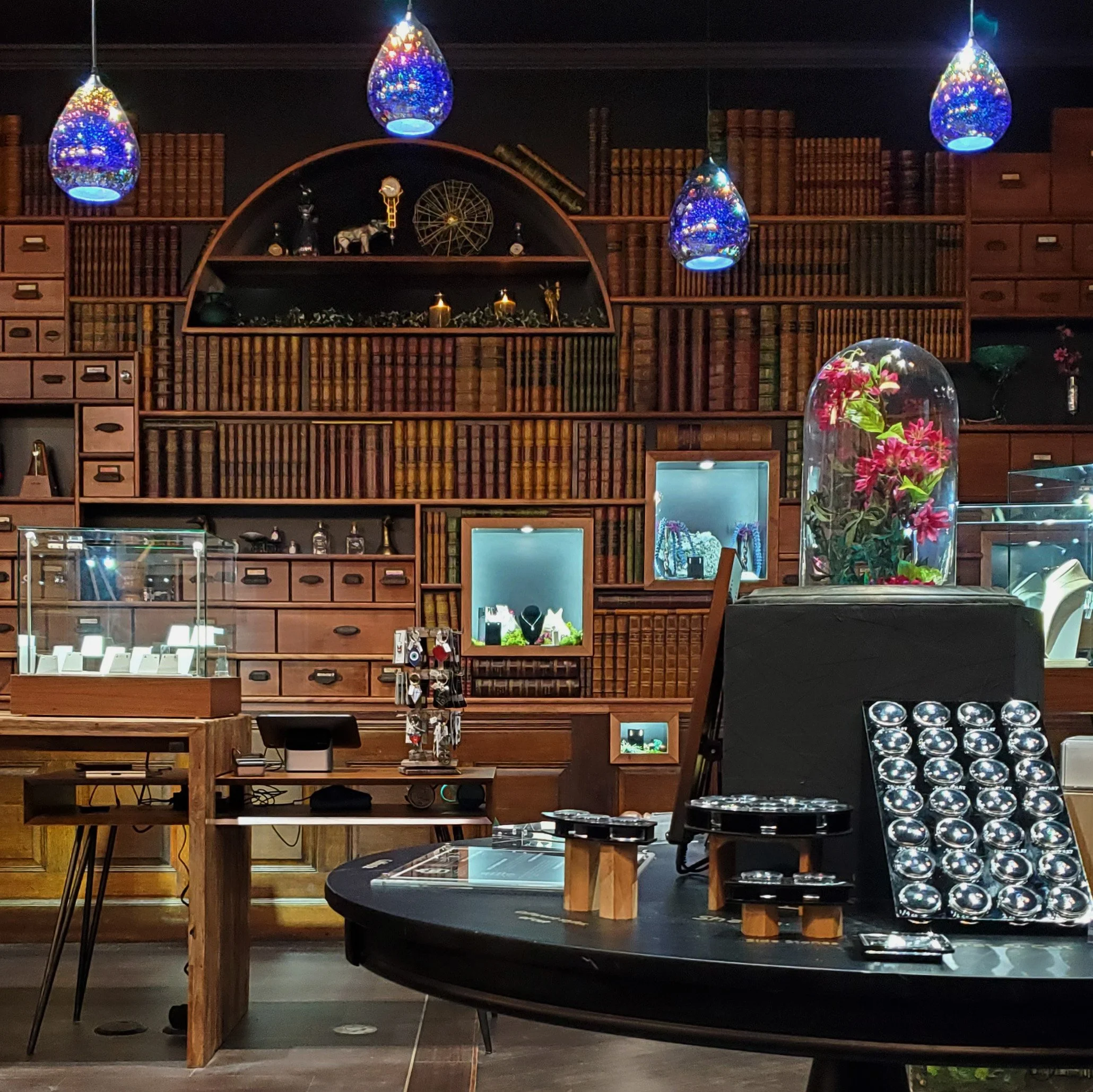 Interior of a store displaying jewelry, with wooden shelving and tables showcasing rings and necklaces. Colorful glass pendant lights hang from the ceiling, and a glass casing contains pink flowers. Shelves are filled with books and decorative items.