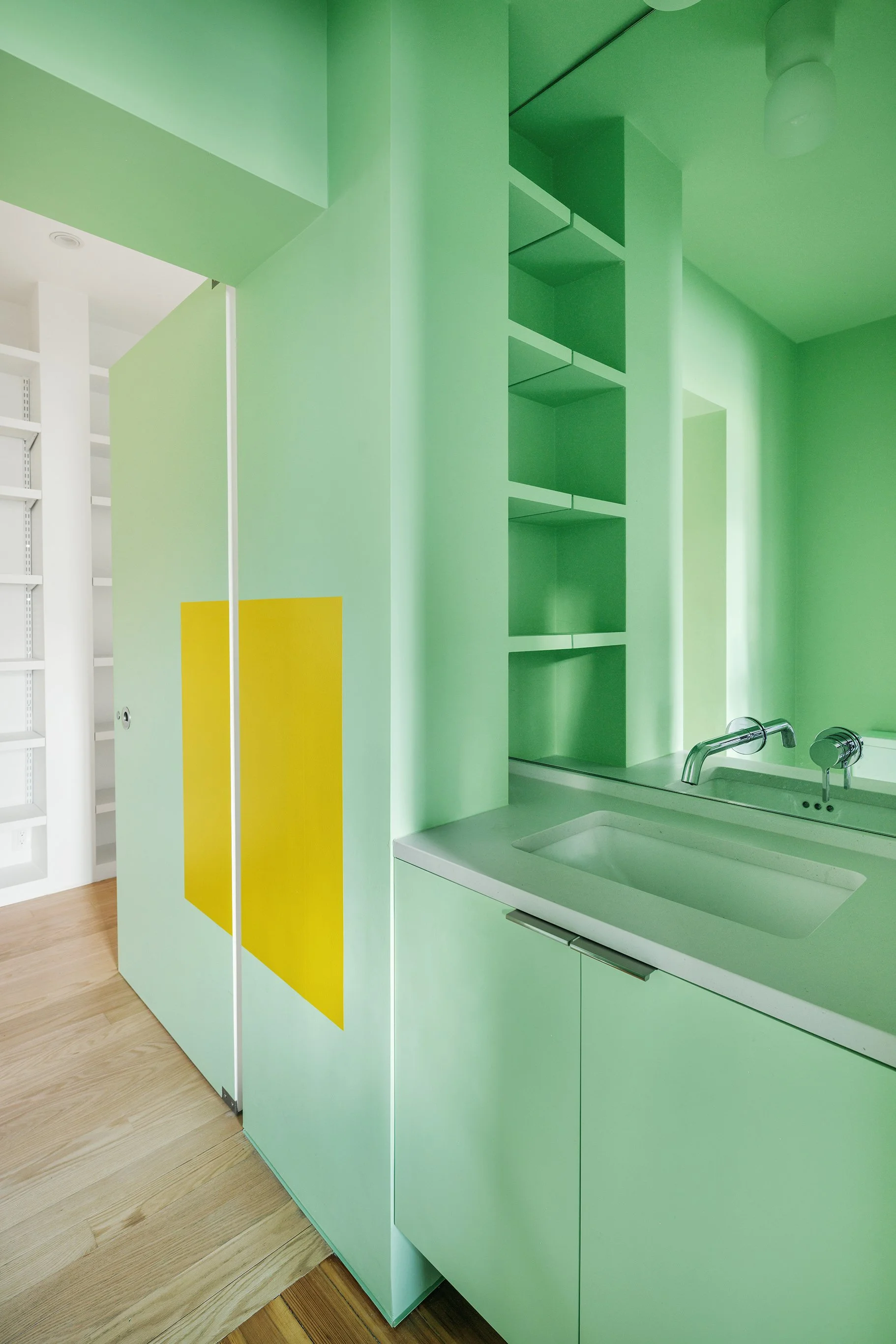 Interior of a modern kitchen with green cabinets, a white sink, and a silver faucet, with light wood flooring.