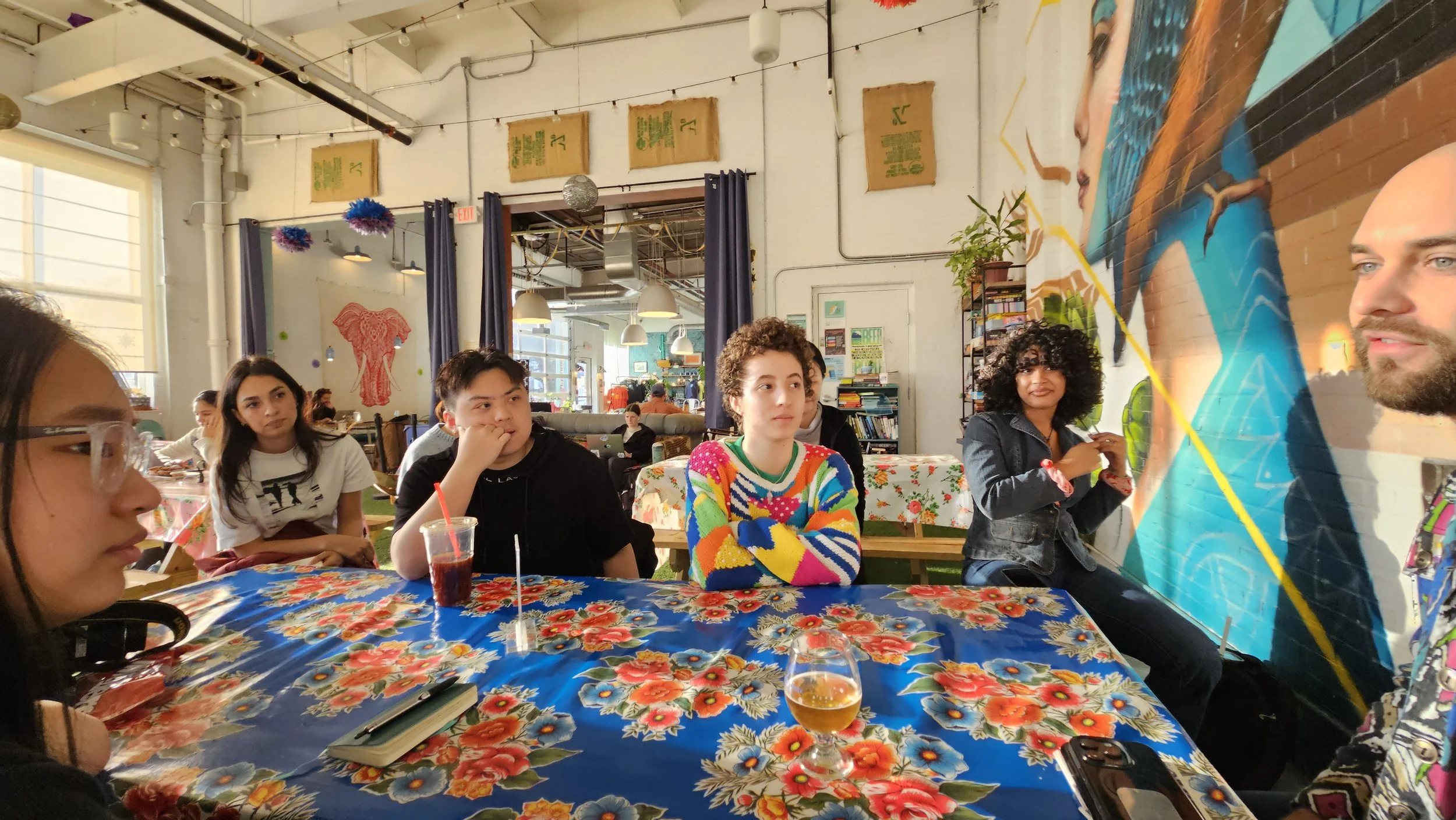 A group of young adults sitting at a table during a gathering or party in a decorated indoor space. The table has a colorful floral tablecloth, and the group appears engaged with a colorful mural of a woman's face on the wall behind them.
