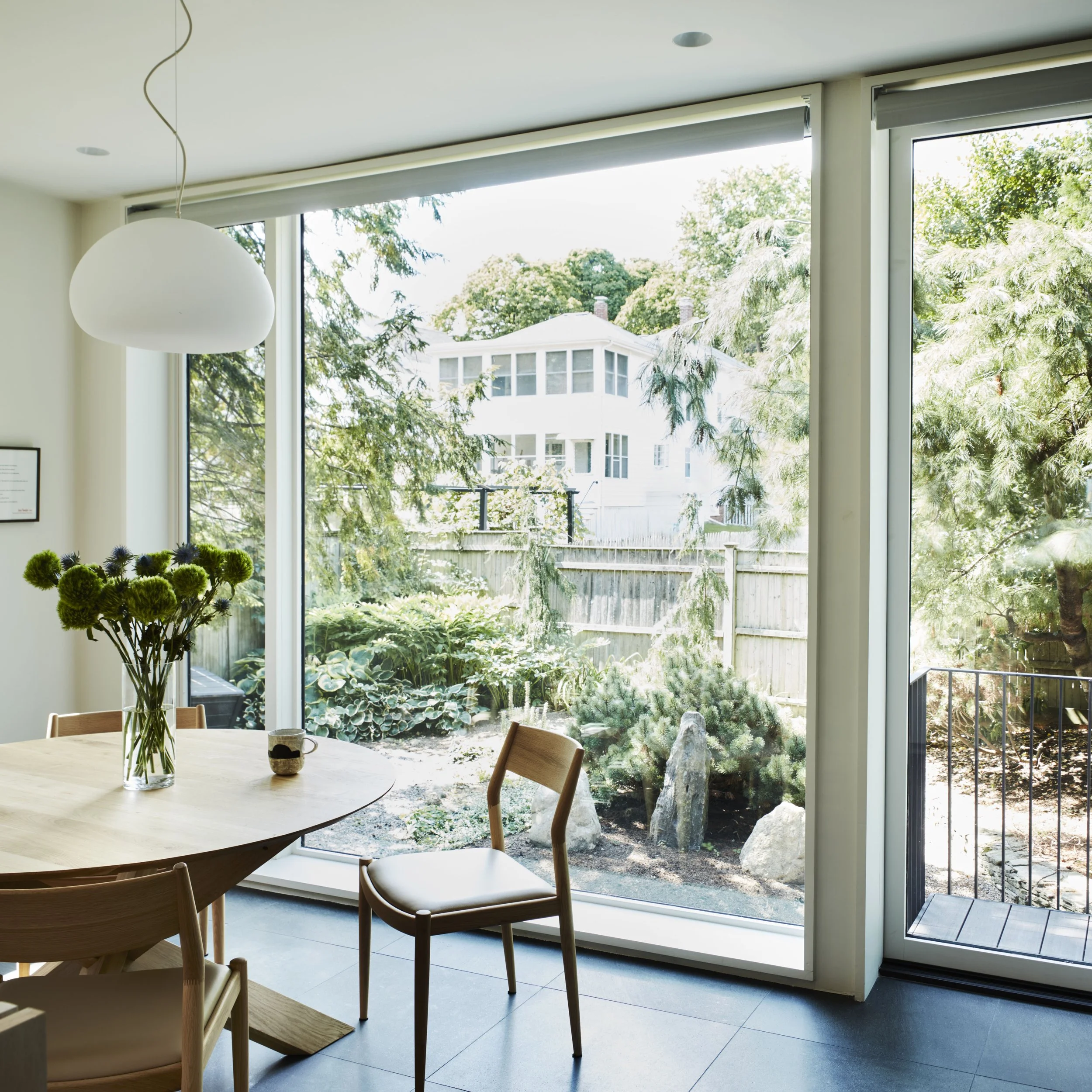 A dining area with a wooden dining table and chairs placed around it. Large glass sliding doors reveal a lush backyard garden with trees and shrubs, allowing plenty of natural light into the room. A hanging white pendant light is visible above the table.