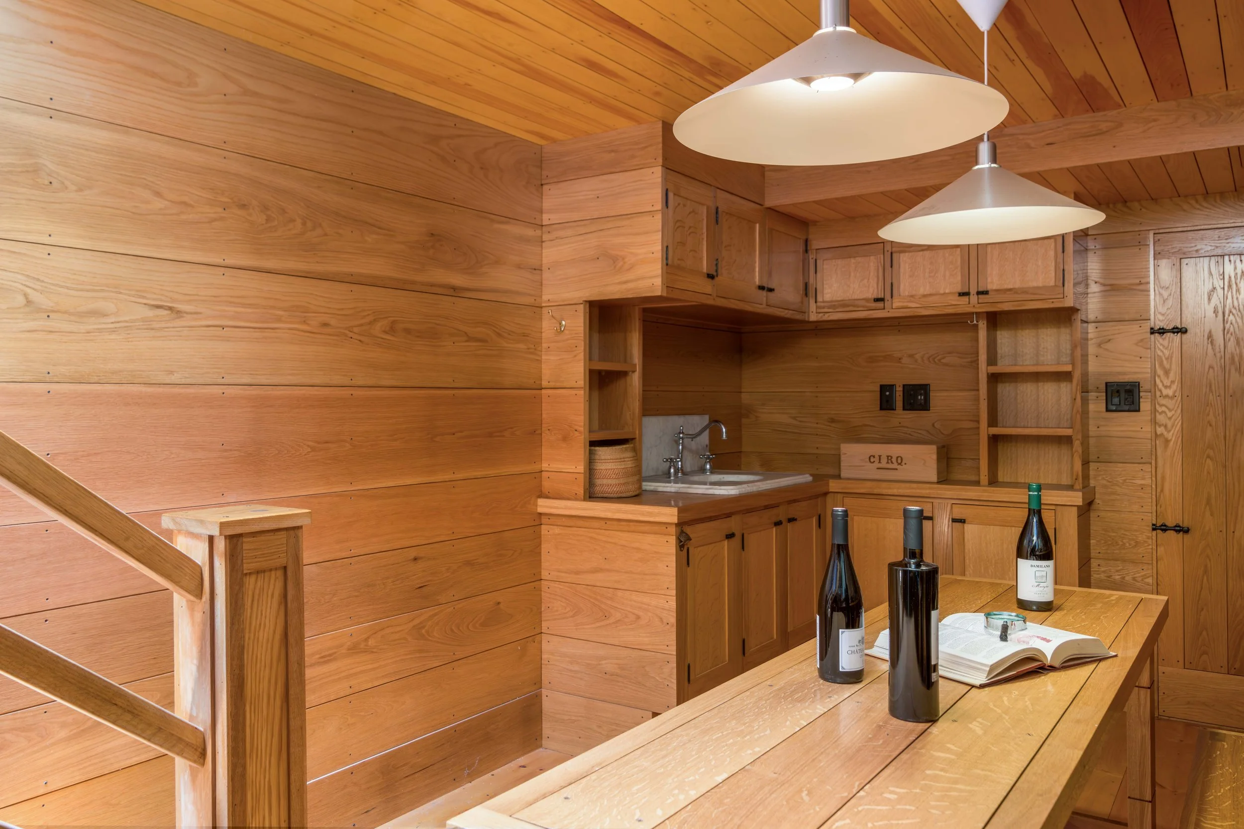 A wooden kitchen with wood-paneled walls and ceiling, featuring a nook with a small sink, countertop, and wooden cabinets. Three white pendant lights hang from the ceiling. On the table, there are three bottles of wine, an open book, and a pair of gl
