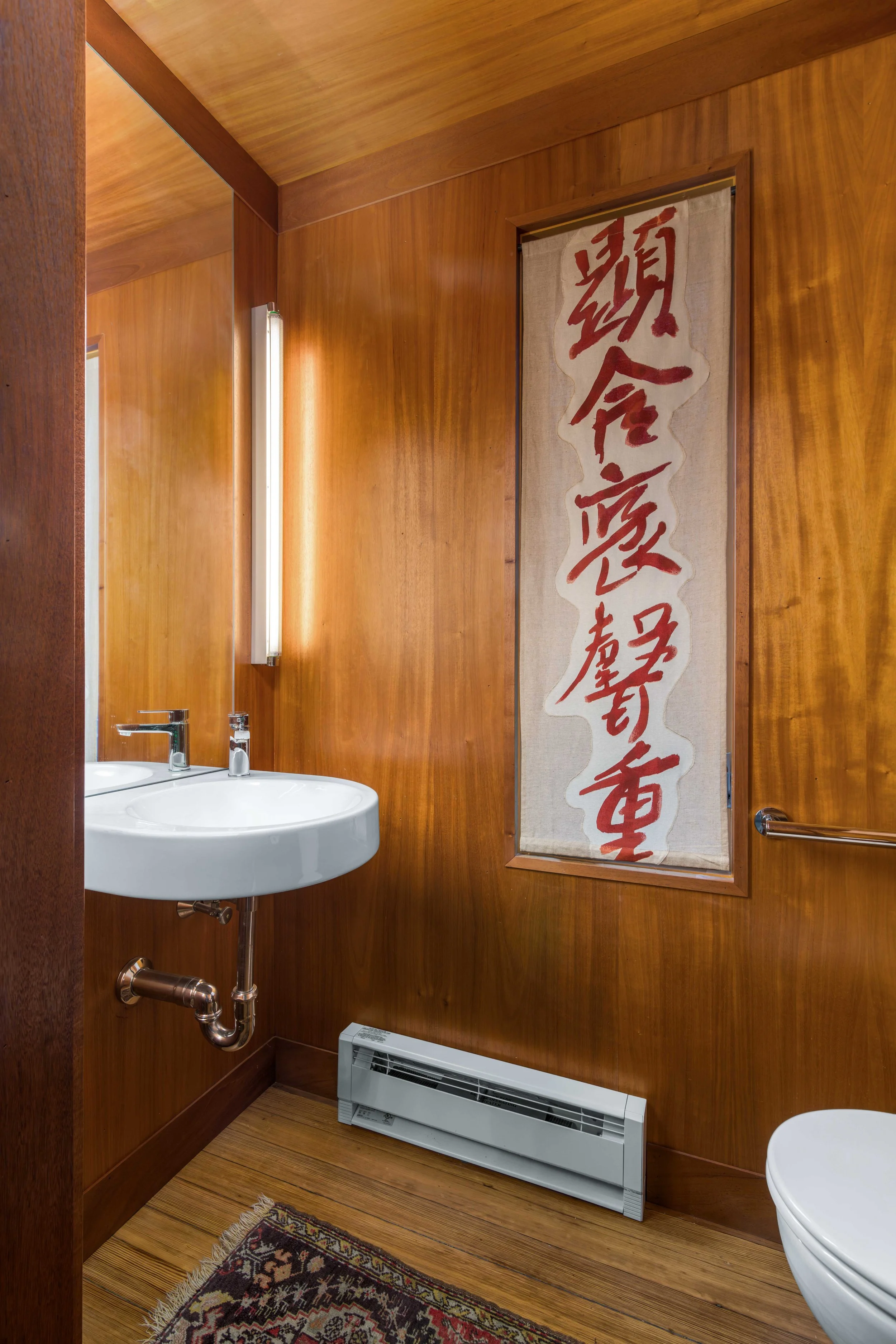 Bathroom with wood-paneled walls, a white sink with a silver faucet, a radiator heater below, and a decorative fabric hanging with Japanese calligraphy.