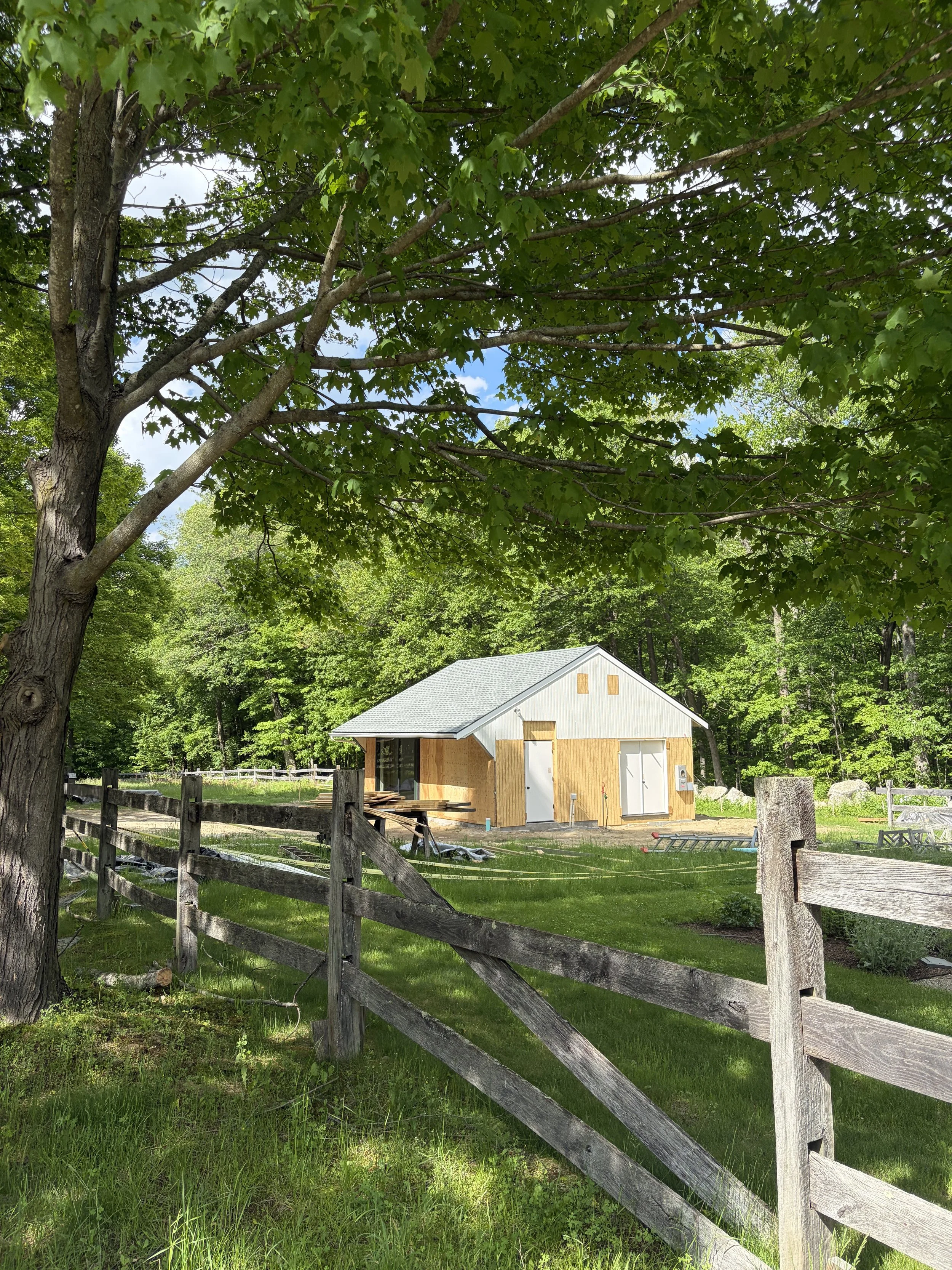 A rural scene featuring a small barn or shed being constructed or renovated, surrounded by a rustic wooden fence, green trees, and lush grass, with a clear blue sky overhead.