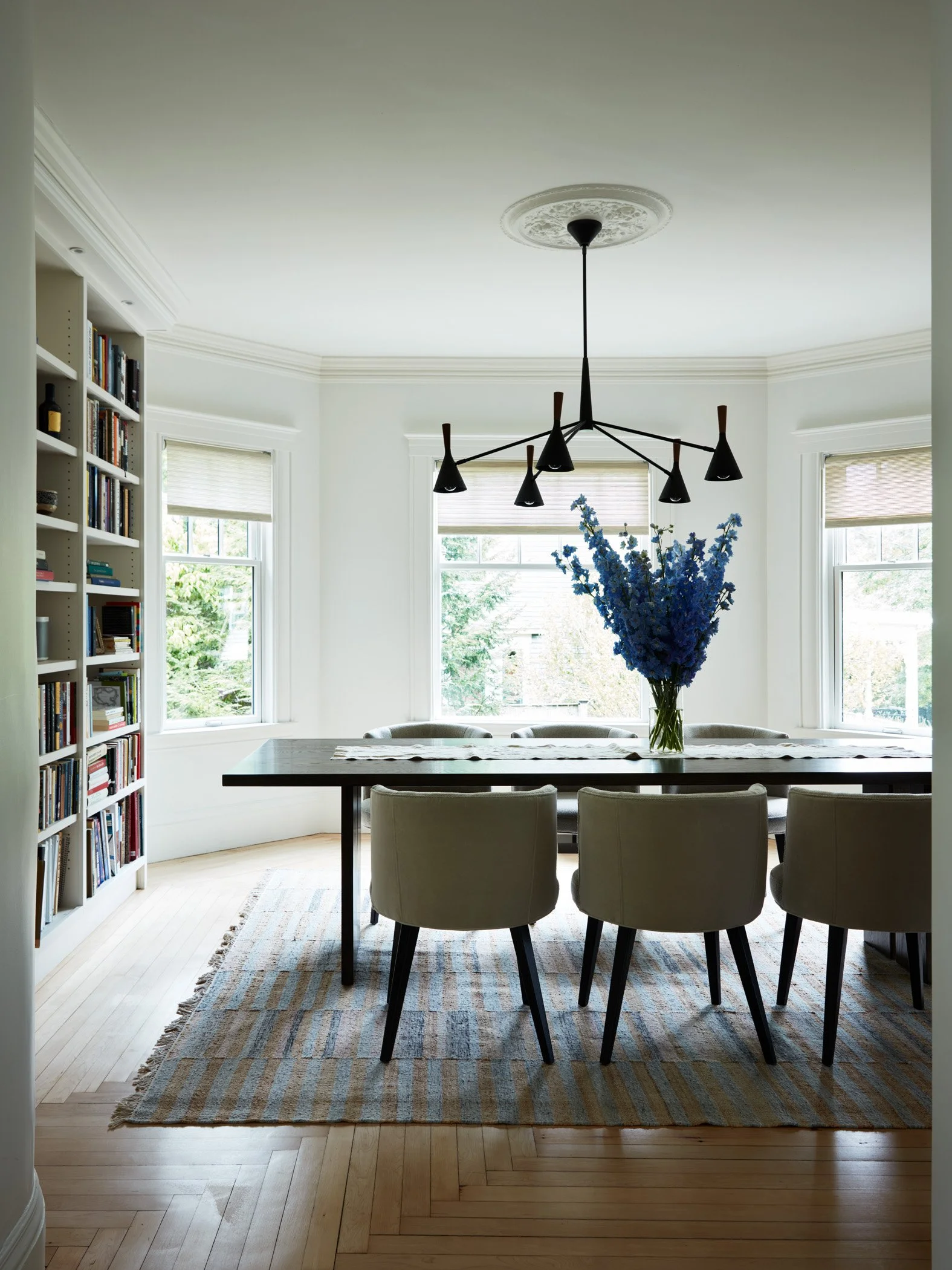 A dining room with a large dark wooden table, six upholstered chairs, and a centerpiece with blue flowers. There are three large windows with blinds, a bookshelf on the left with books and decorative items, a patterned rug under the table, and a mode
