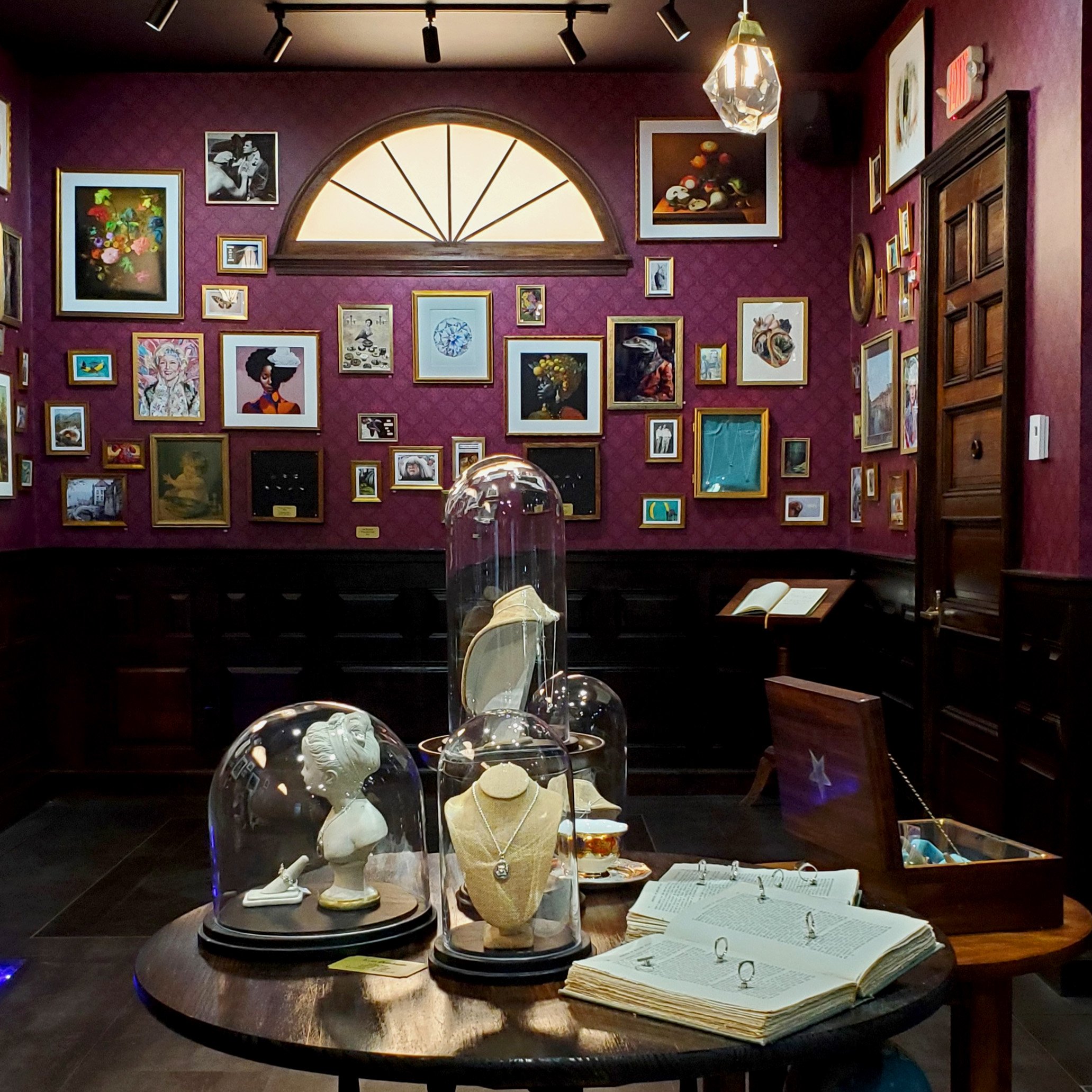A gallery wall with various framed artwork, including photographs, paintings, and illustrations, in a room with dark red walls and a semi-circular window. In the foreground, glass domed display cases hold white sculptures, and open books are placed o