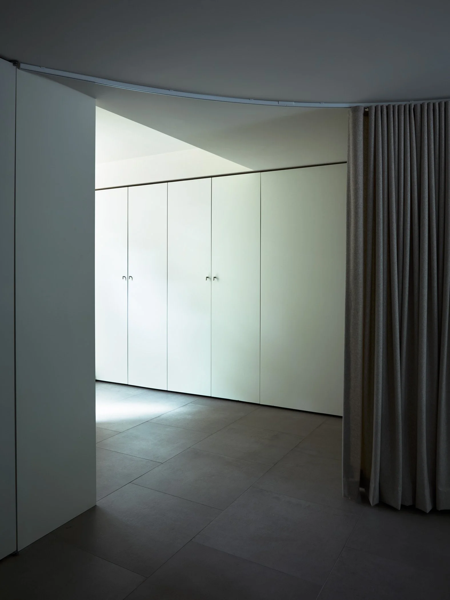 A modern interior space with a white wardrobe, a curtain on the right side, a sliding door on the left, and a high ceiling with a unique curved light fixture.