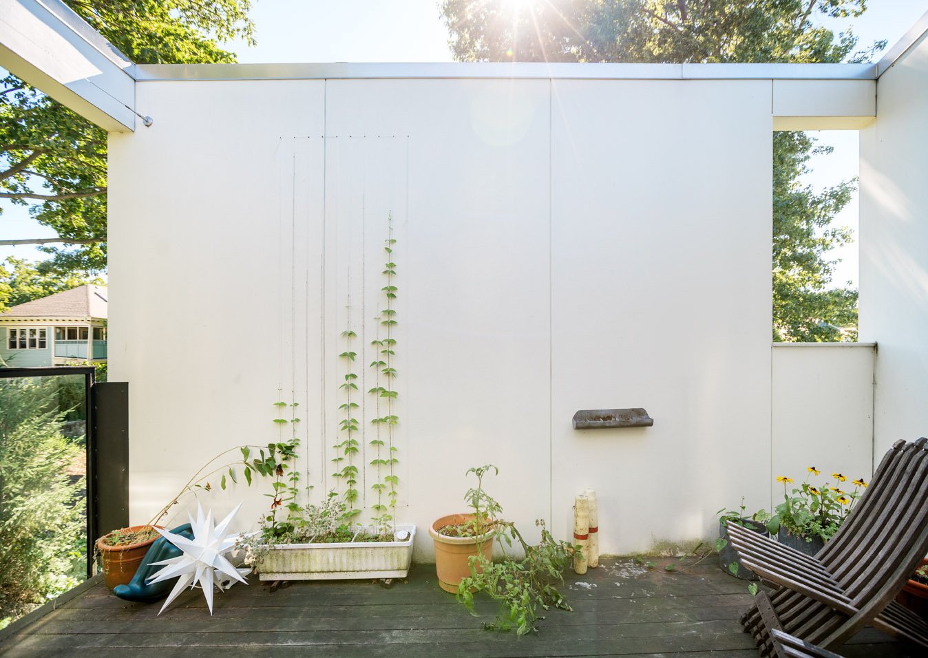 A small outdoor balcony with potted plants, a white decorative star, and a striped wooden chair, with sunlight shining through trees in the background.