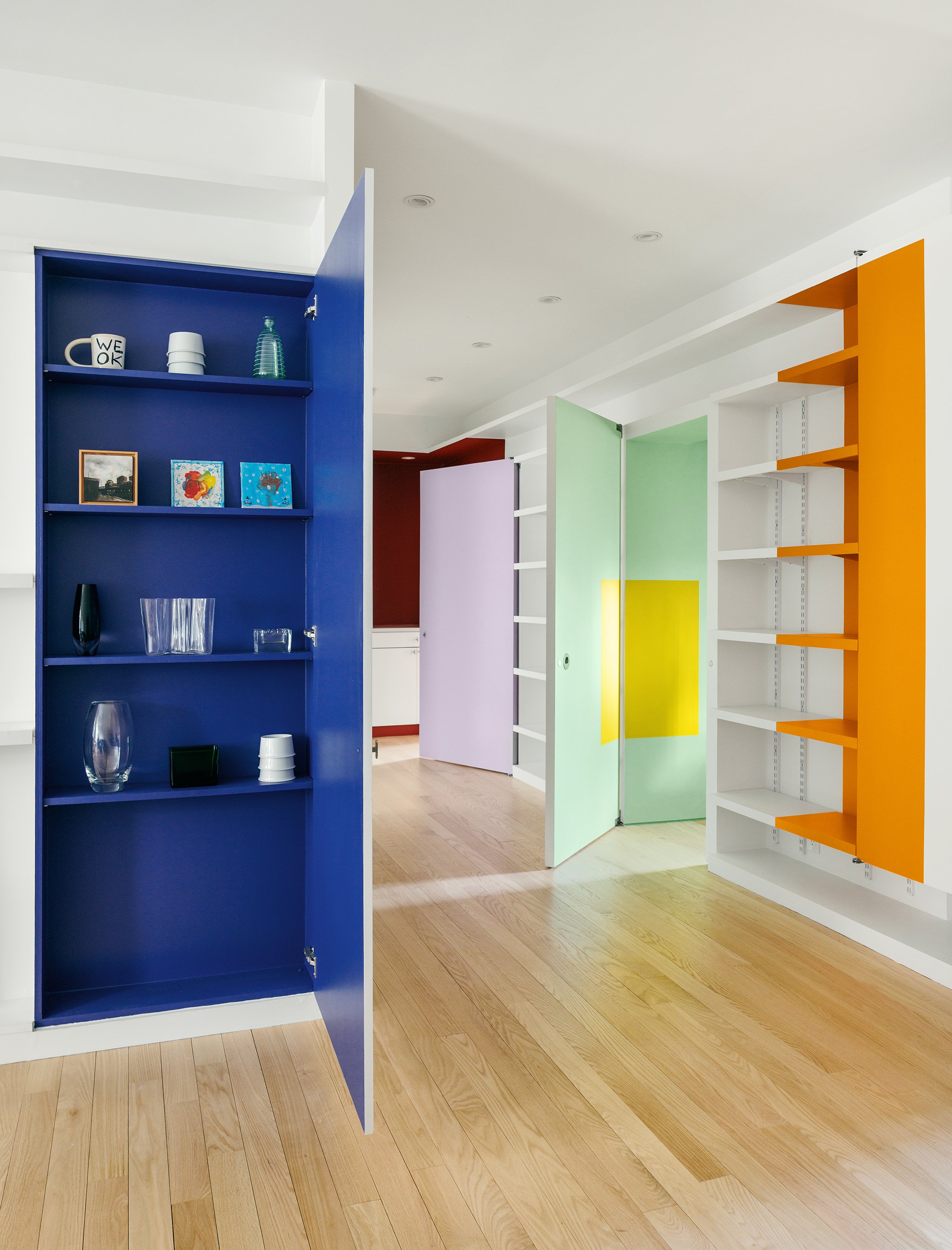 Colorful room with open cabinets and partitions painted in blue, green, purple, yellow, and orange. The room has wooden flooring and contains various decorative items and artwork on the shelves.