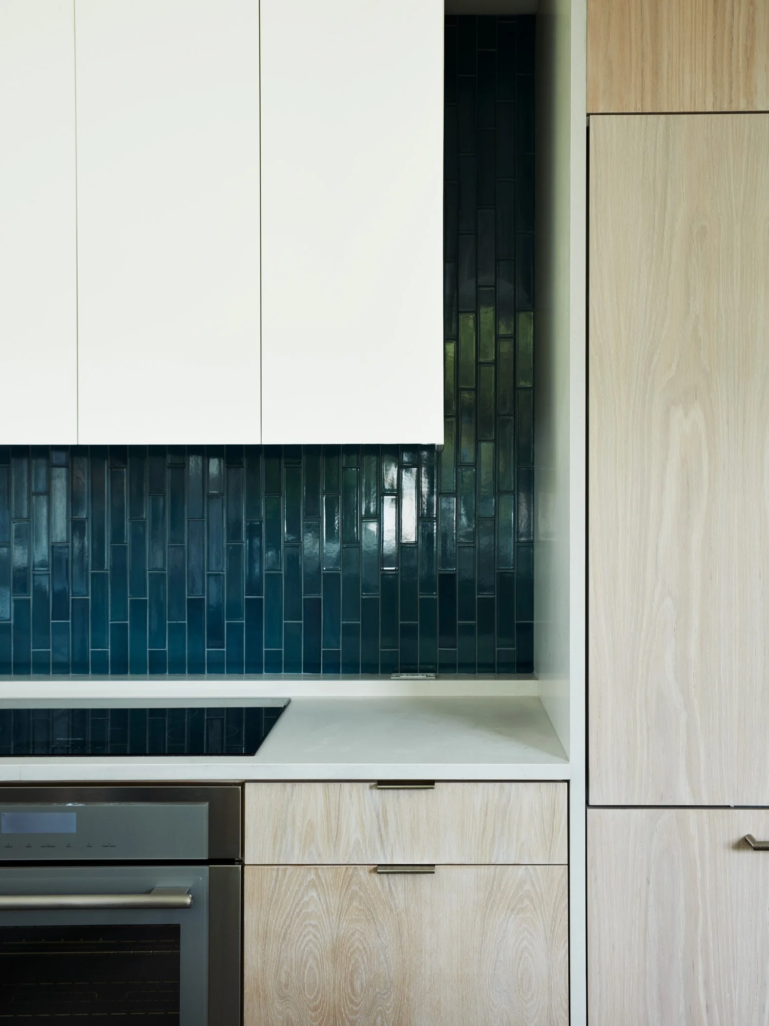 Close-up view of a modern kitchen cabinet wall with a black tile backsplash, featuring a built-in oven and light wood cabinets.