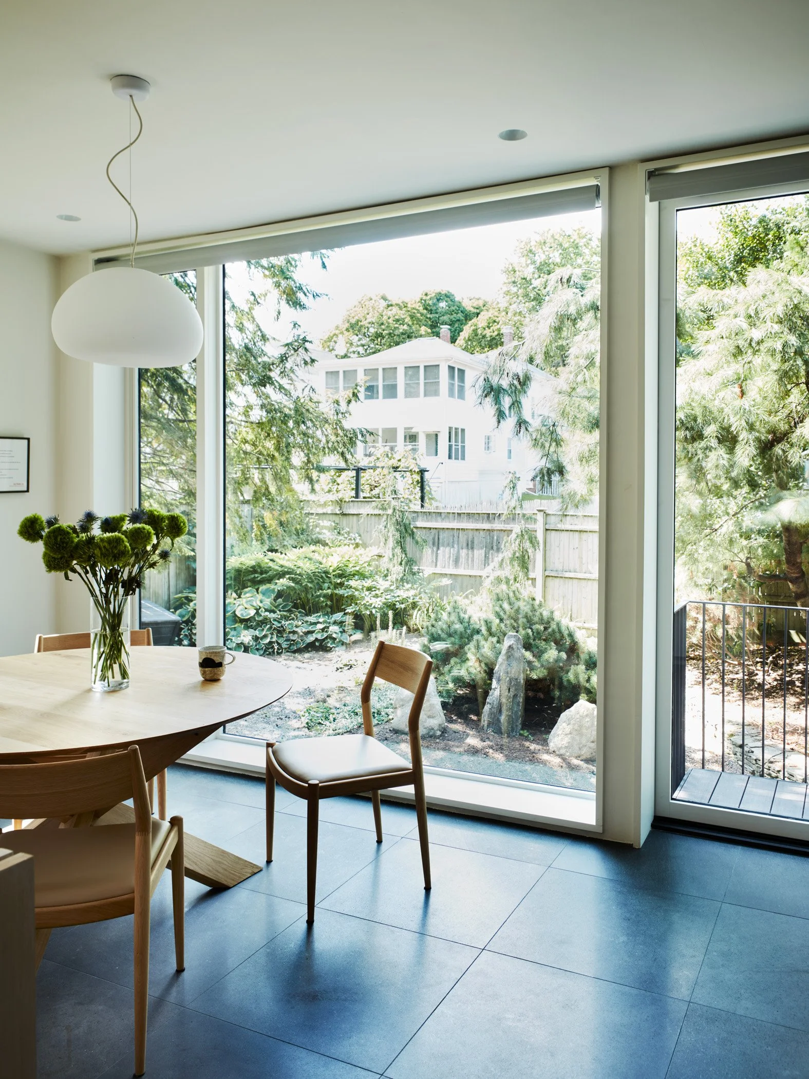 Interior of a modern dining area with a round wooden table, light-colored chairs, a white pendant lamp, and large windows revealing a garden with trees and a white house in the background.