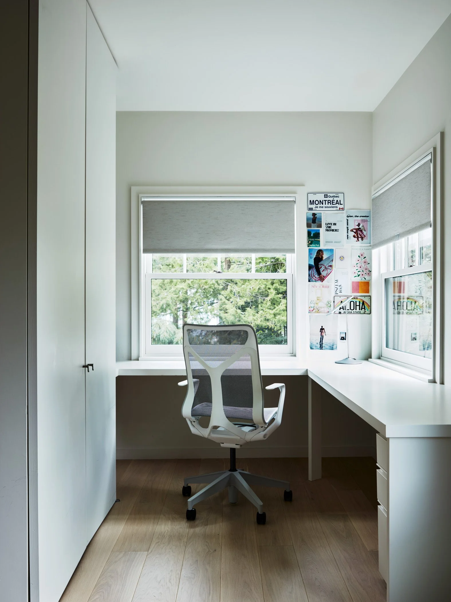 A modern, minimalist home office with a white desk, an ergonomic office chair, large windows with shades, and a wall decorated with posters and a sign that says Montreal.