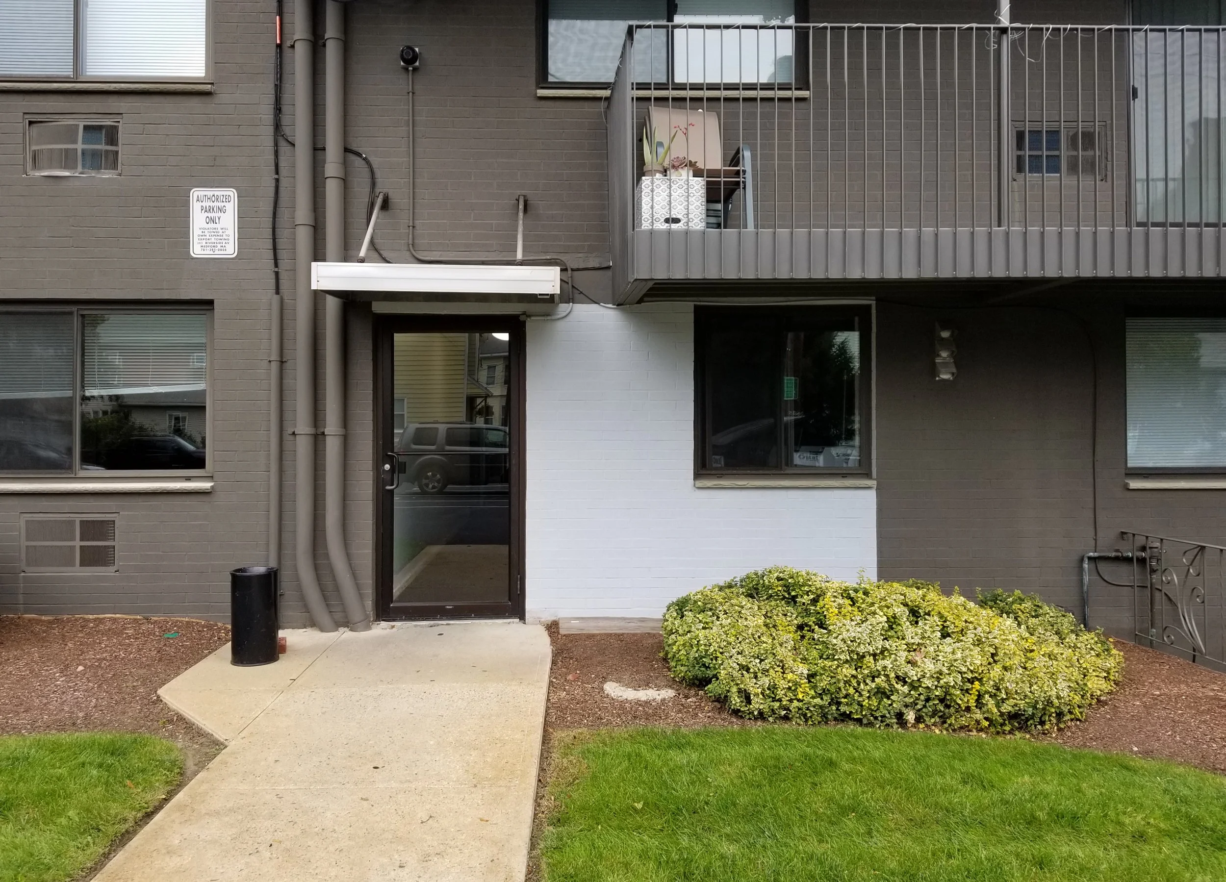 Exterior view of a modern apartment building with a small porch area, gray brick facade, and a balcony with plants. There is a sign indicating authorized parking only, and a small landscaped area with bushes and grass.