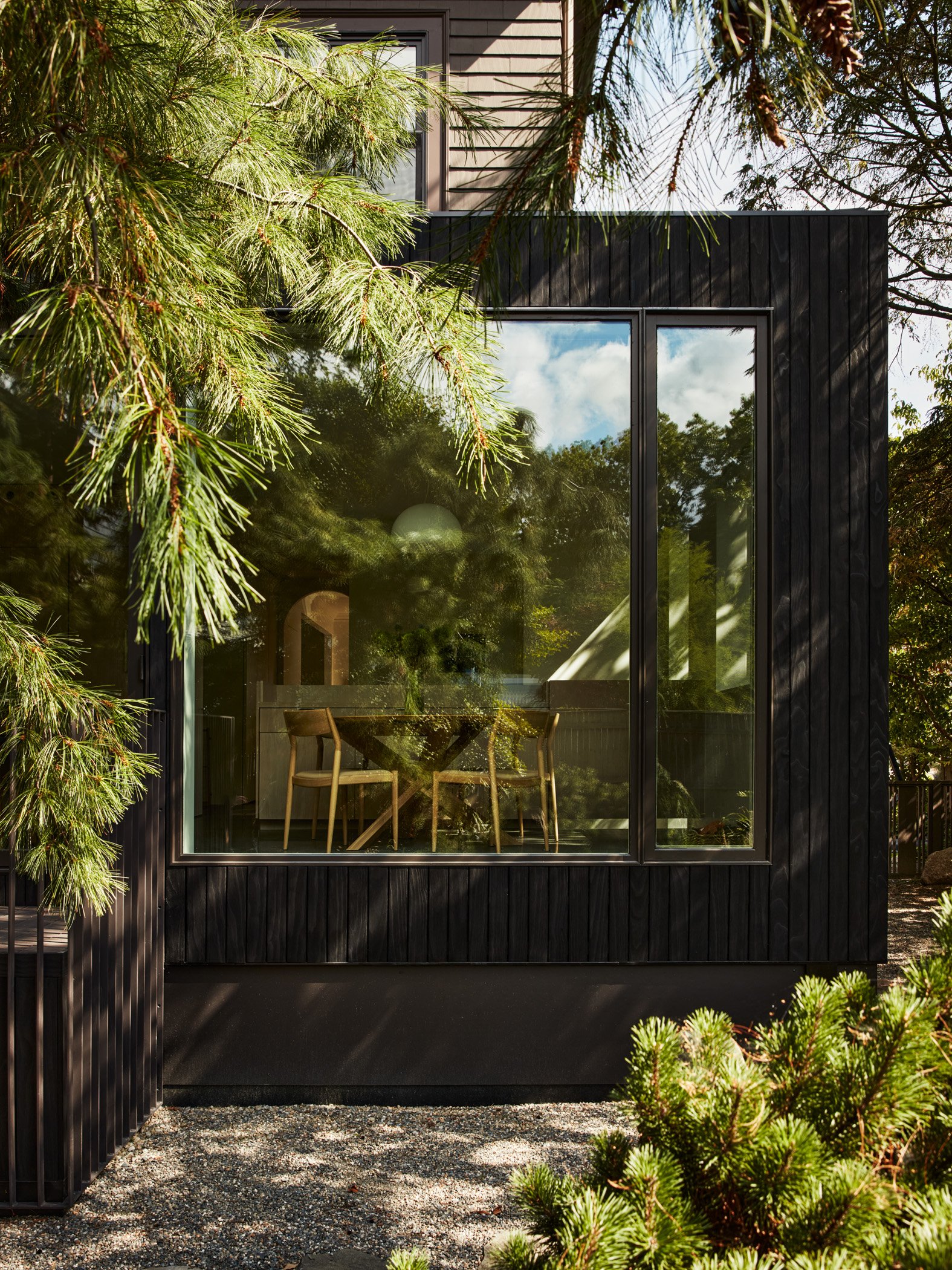 Modern black house with large glass windows, surrounded by green trees and shrubs, interior with a table and chairs visible through the glass.