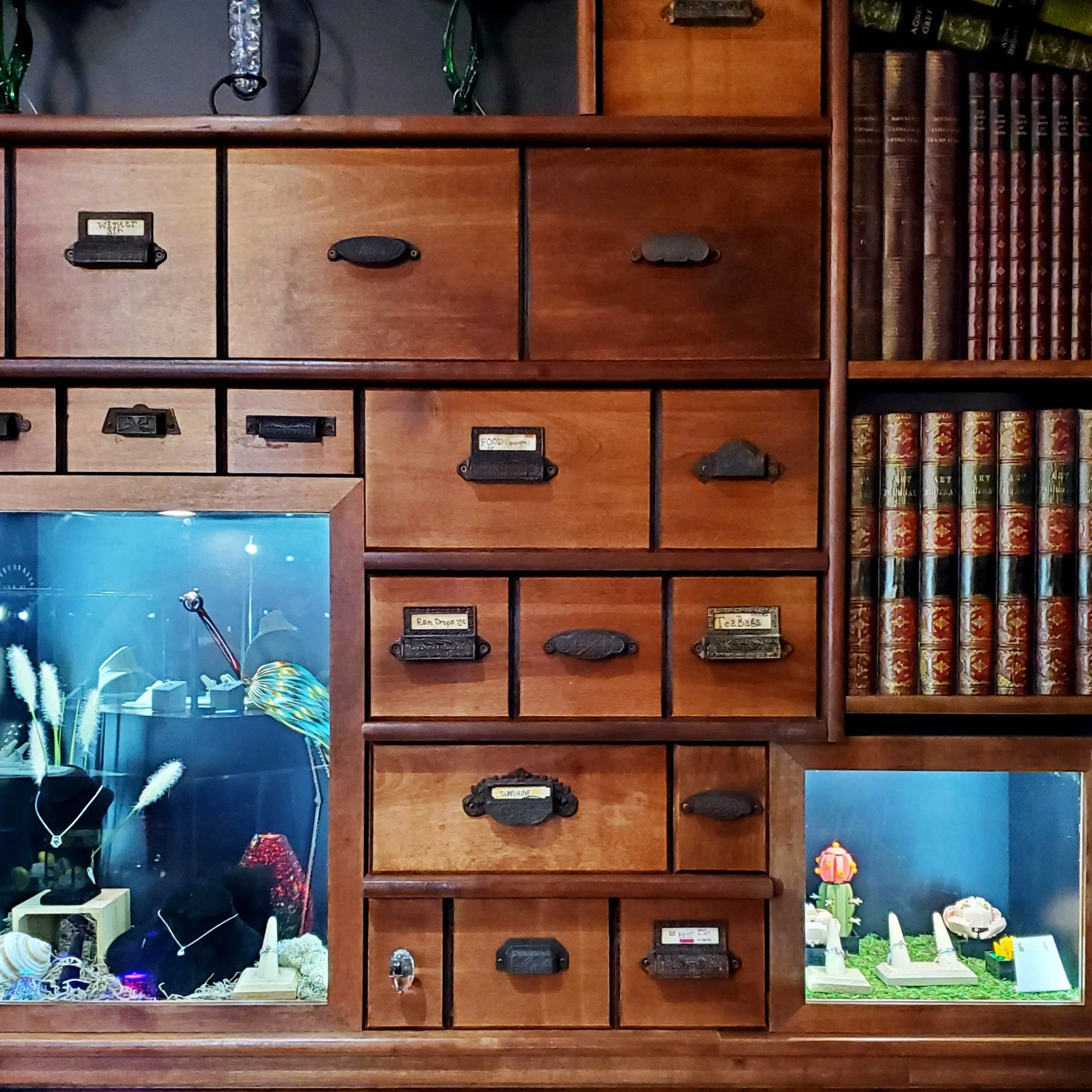 A wooden shelf with multiple compartments containing decorative items and books. The top left compartment has a small aquarium with fish and seaweed decorations. The second row middle compartment has a label that says 'Rain Drops 50¢'. The bottom rig