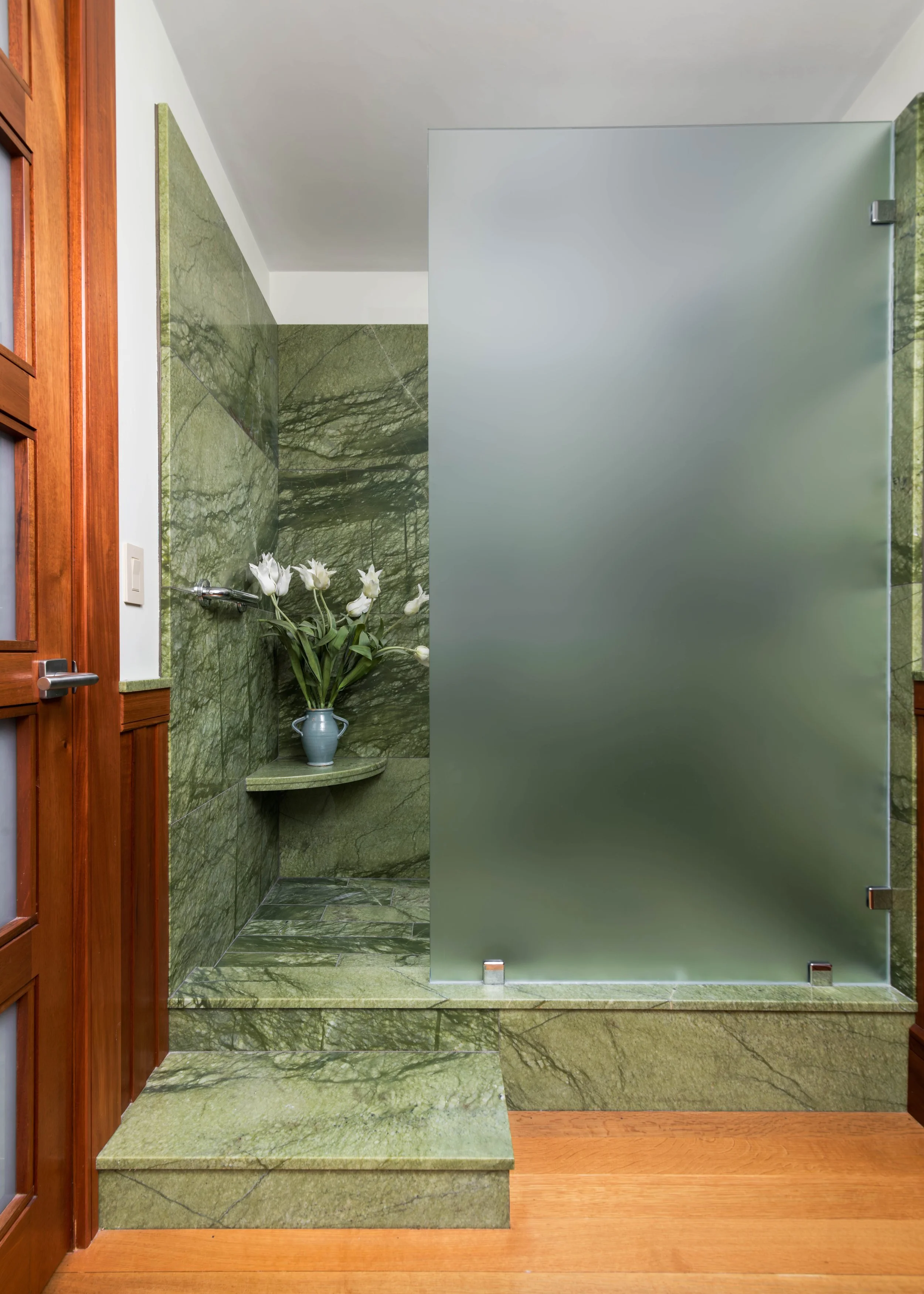 Bathroom with green marble tiles, a silver frosted glass shower door, a wooden frame door on the left, and a blue vase with white flowers on a green marble ledge.