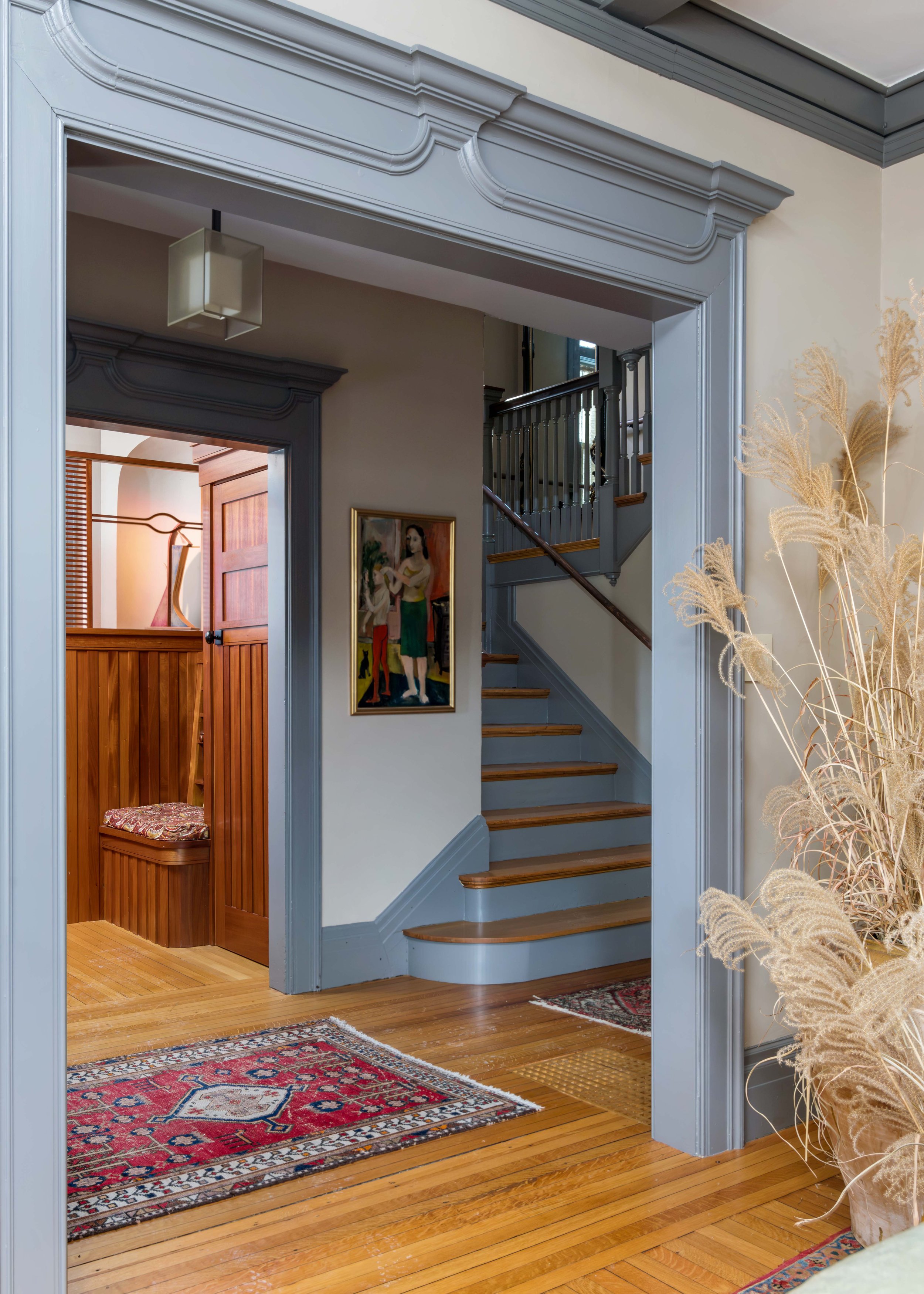 Interior view of a home featuring a staircase with wood steps and a gray railing, a decorative framed painting of a woman, a multicolored area rug, and a doorway with a wooden door, all inside a traditional style house with hardwood floors and detail