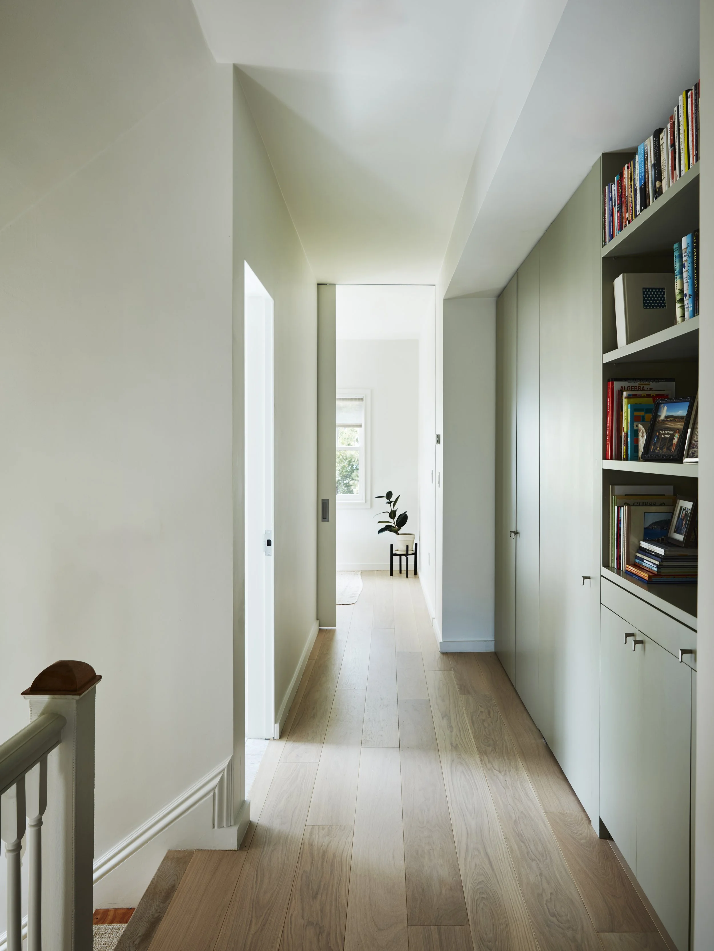 A bright hallway with light wooden flooring, white walls, and built-in shelves filled with books on the right side. Natural light coming through a window at the end of the hall illuminates a potted plant and a small stool.