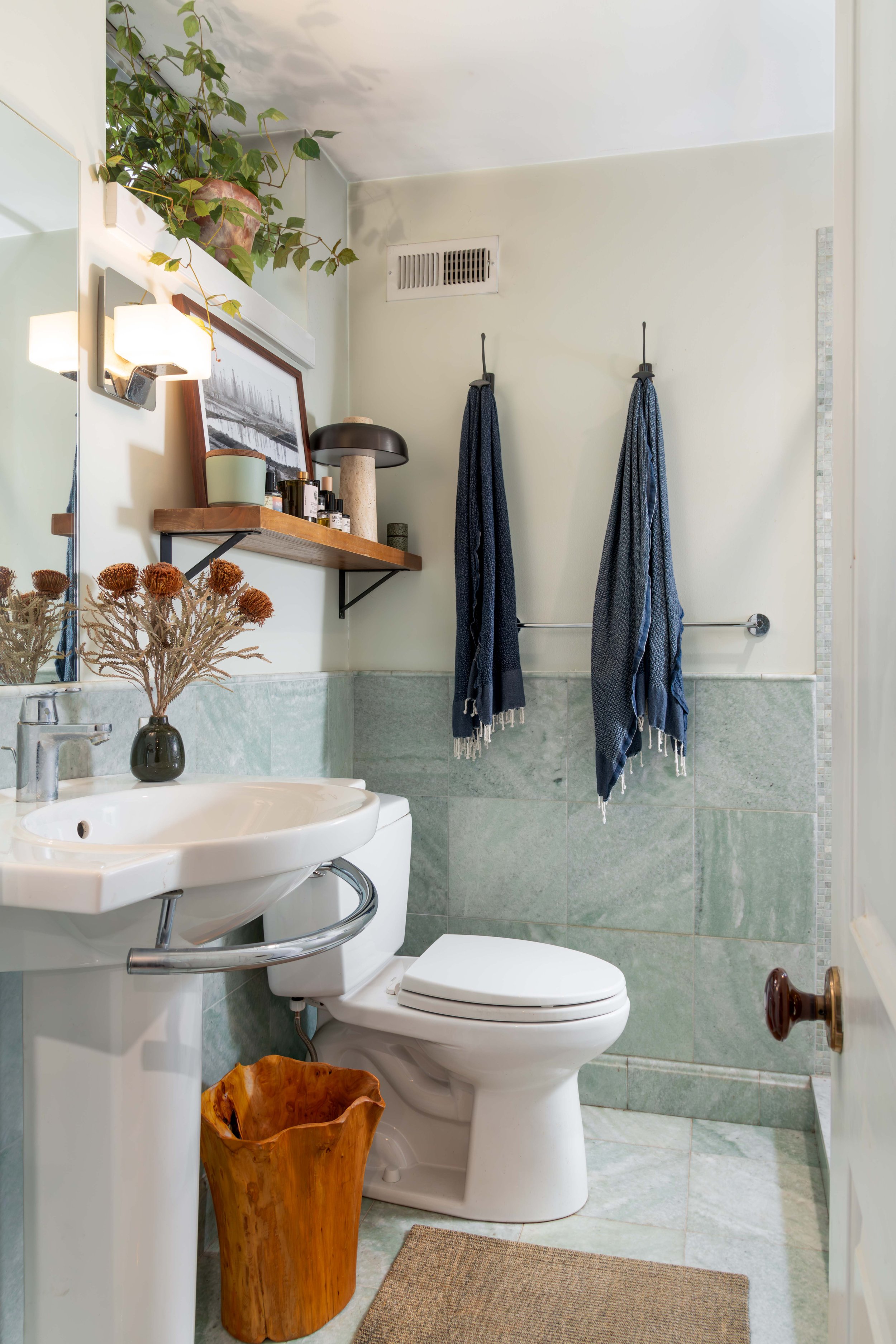 A small bathroom with a white sink, a toilet, and a mirror reflecting a toiletry shelf. The shelf holds various bottles, a black table lamp, and a picture with a wooden frame. Hanging on the wall are two dark blue towels on hooks. There is a potted p