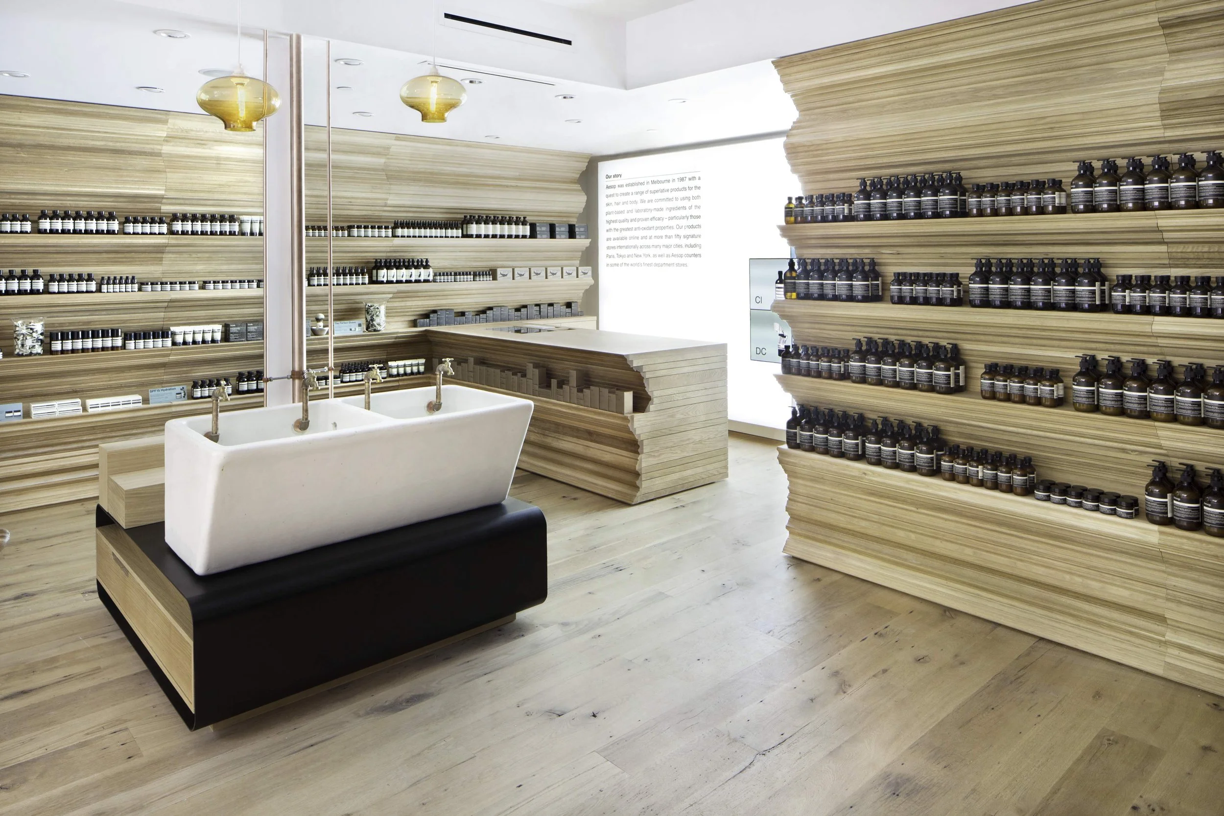 Interior of an AESOP store with wooden shelves stocked with small bottles, a large, white ceramic sink with copper pipes, and warm pendant lighting.