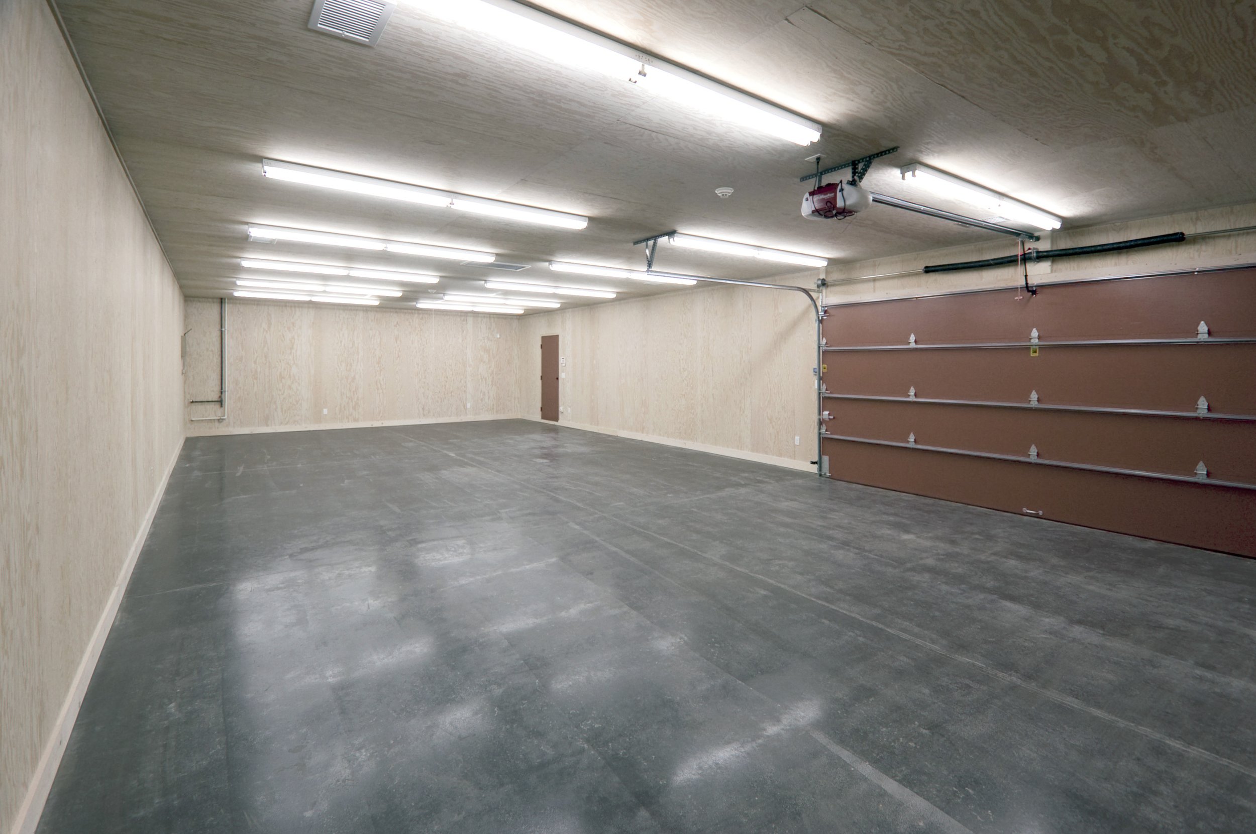 Empty garage with fluorescent lights, beige walls, concrete floor, brown garage door, and a door at the back.