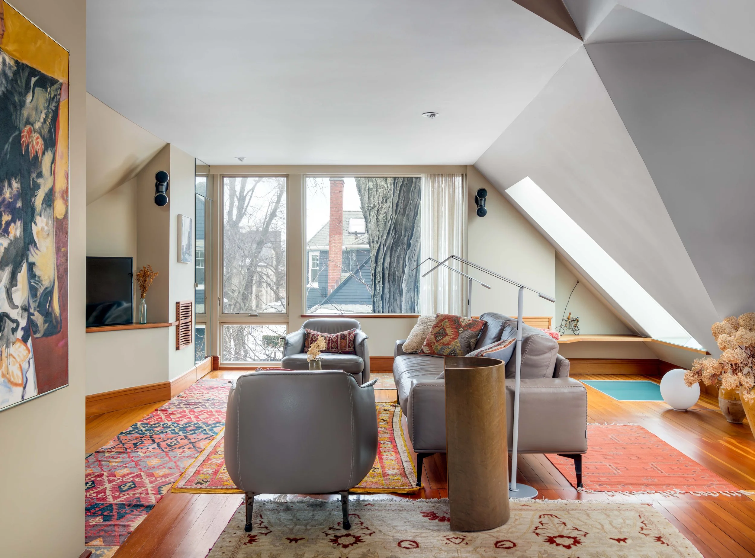 A living room with large windows showing a wintery outdoor scene. The room has a sloped ceiling with built-in speakers, a colorful area rug, a gray sofa with patterned cushions, a black leather armchair, decorative vases, and a colorful abstract pain