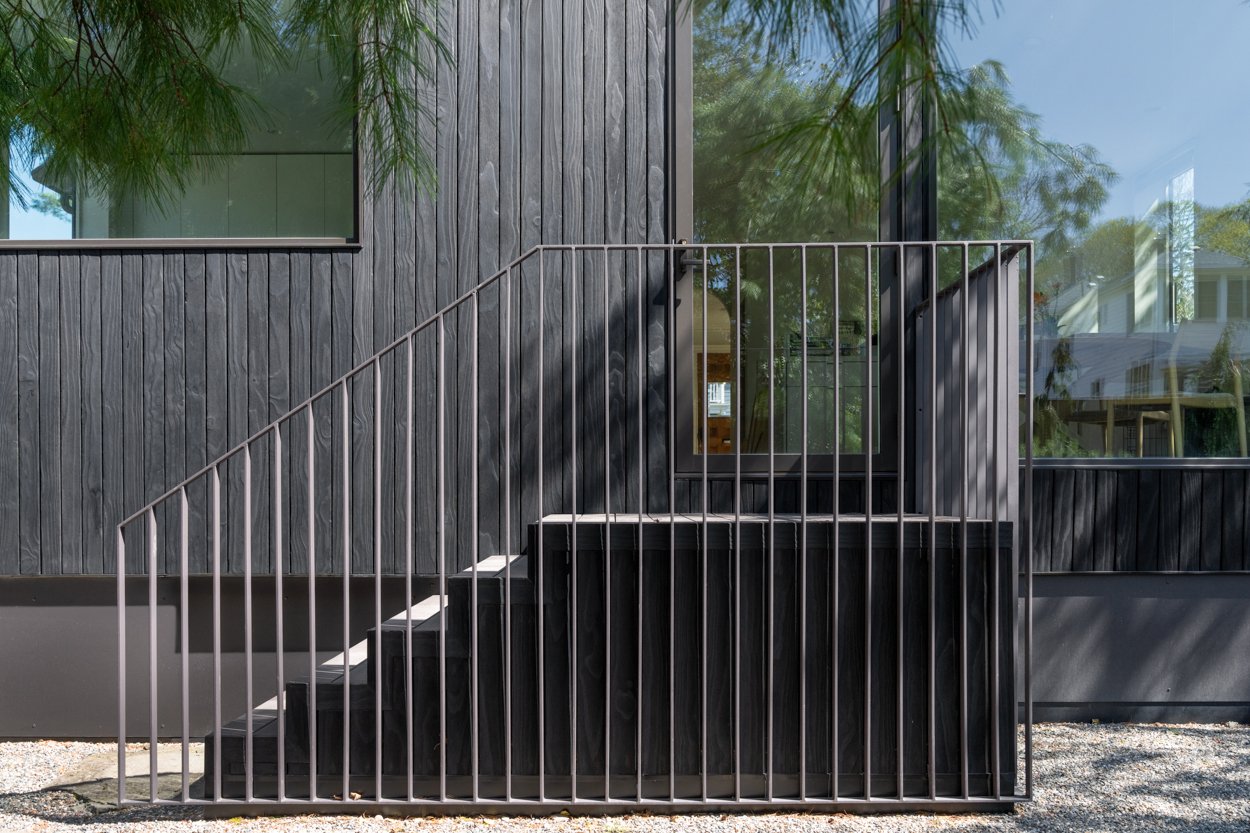 Black exterior staircase with metal railing leading to a door with glass window, attached to a modern gray building with large windows and wooden paneling. Green trees reflect in the windows.