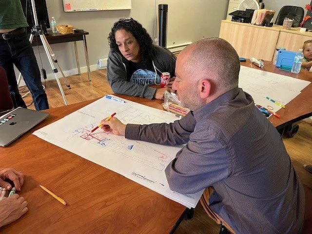 Two adults sitting at a table looking at a large sheet of paper with drawings and notes. One person is writing on it with a red marker, and the woman is holding a soda can. There is a laptop and a few other objects on the table, with a child in the b