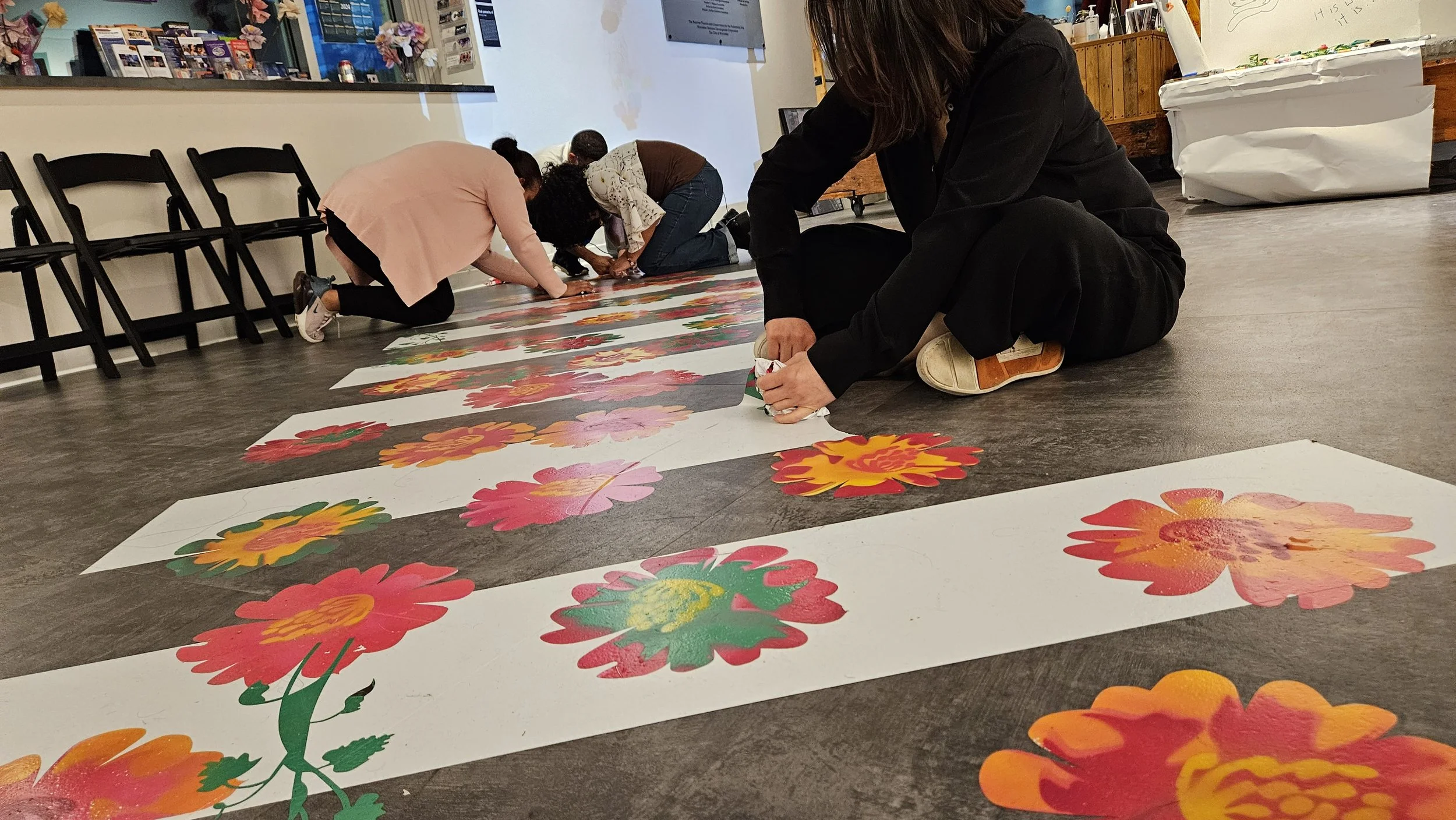 People creating large colorful flower artwork on floor in a public space.