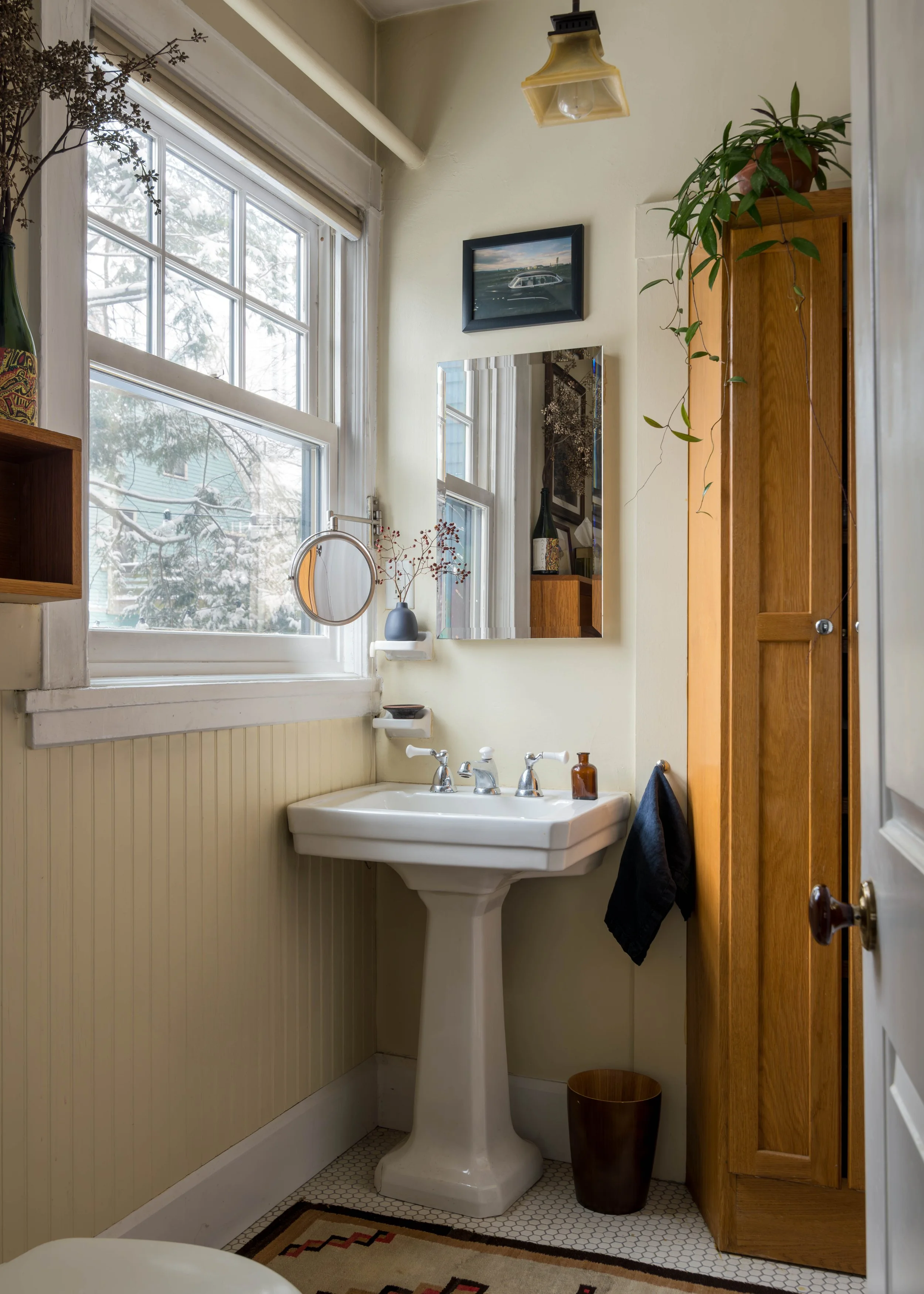 A vintage bathroom corner with a white pedestal sink, a large window showing snow outside, a mirror above the sink, a framed picture of a car, a potted plant on a wooden cabinet, a small wall shelf with a vase, a brown trash can, a towel hanging, and