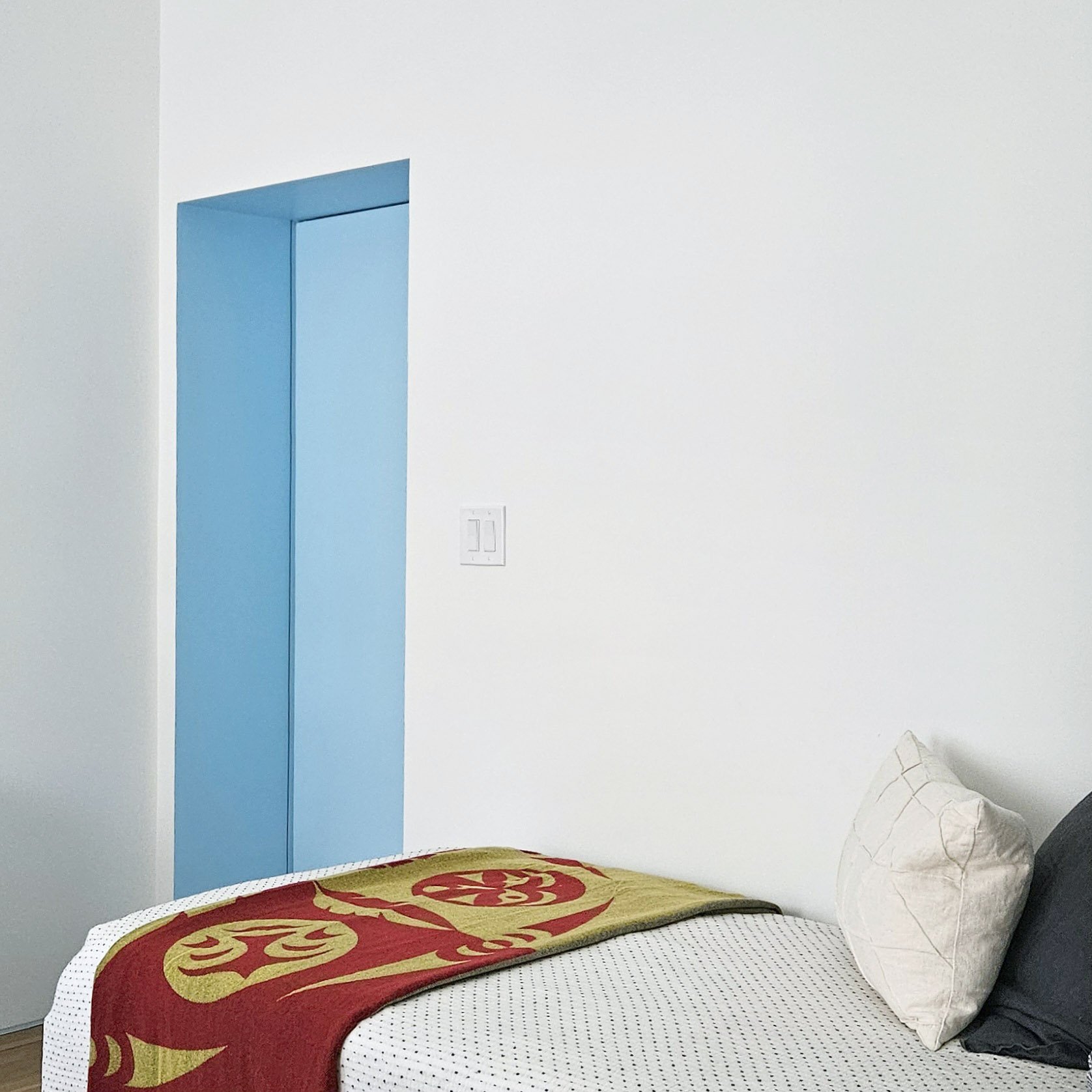 A bedroom with white walls and a blue closet door, a bed with a white mattress and a decorative red and gold blanket, a white pillow, and a black and white pillow. There is a white electrical switch on the wall and part of a beige pillow with a grid 