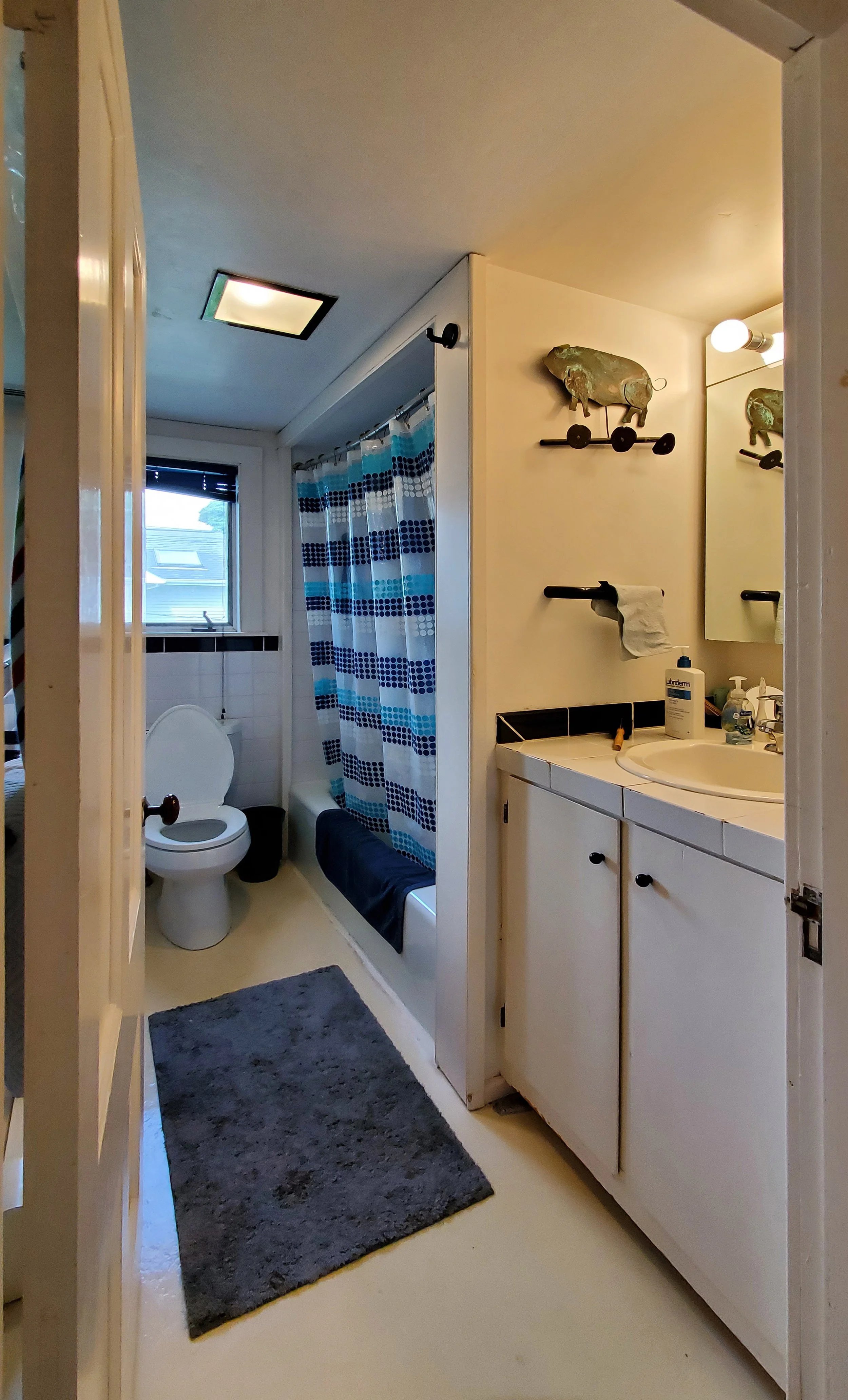 A small bathroom with a toilet, window, shower with blue and white curtains, gray rug, white sink cabinet with toiletries, and wall decorations including a pig sculpture on a shelf and a mirror.